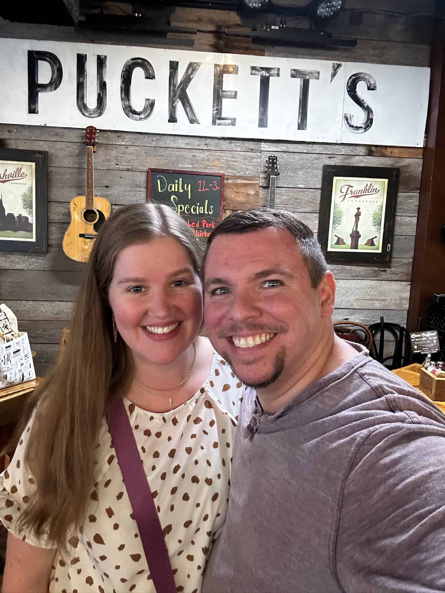 A smiling man and woman snap a selfie inside a Nashville gem—a rustic restaurant with wooden walls and a "Puckett's" sign above them. Guitars and framed pictures adorn the cozy space, capturing the city’s musical spirit.