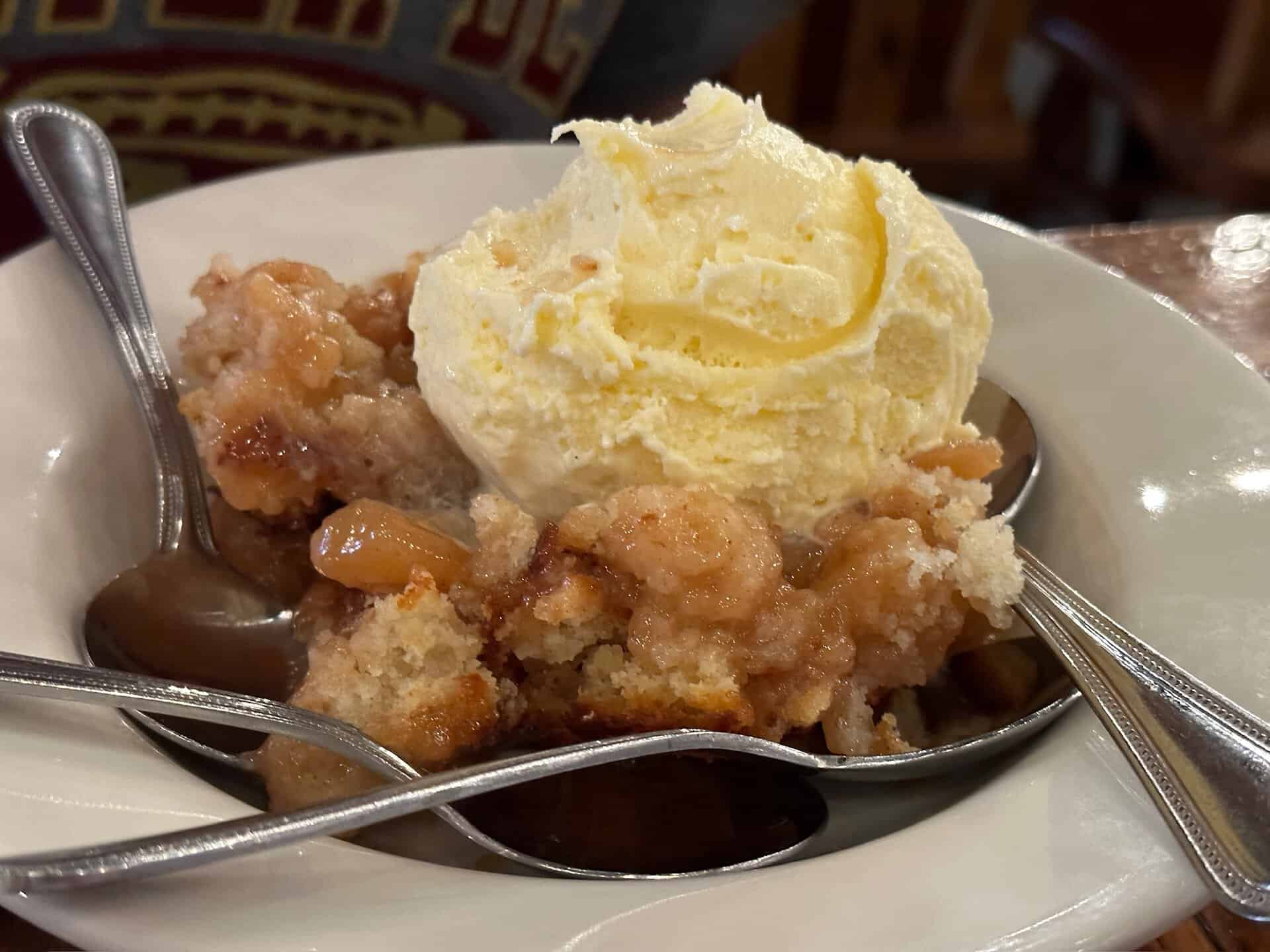 A bowl of apple cobbler, Nashville-style, crowned with a scoop of vanilla ice cream awaits, accompanied by four spoons.