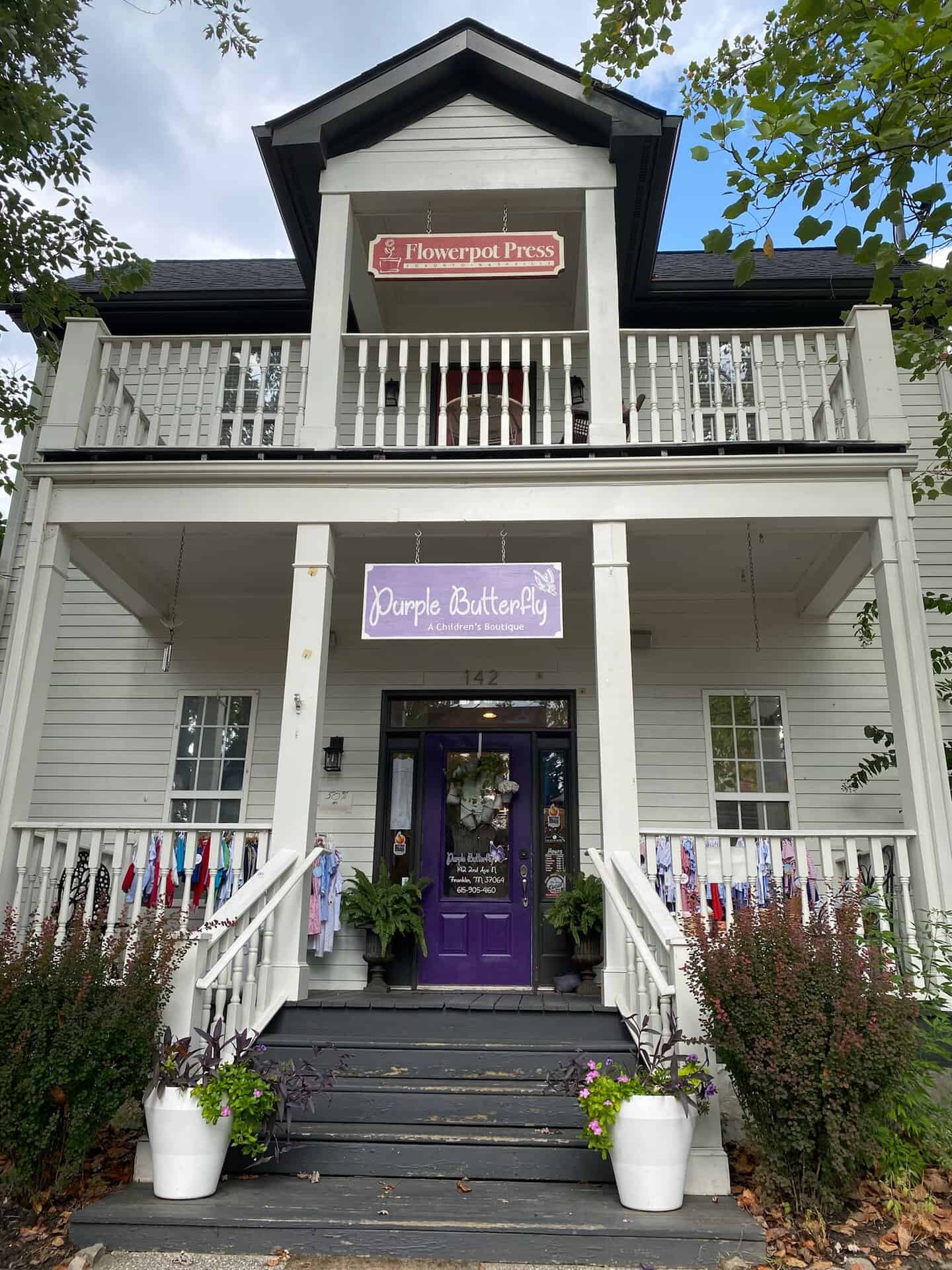 A charming two-story building in Nashville features signs reading "Flowerpot Press" and "Purple Butterfly." The entrance, marked by a purple door and lush plants, welcomes visitors with stylish clothing displayed on rails outside.
