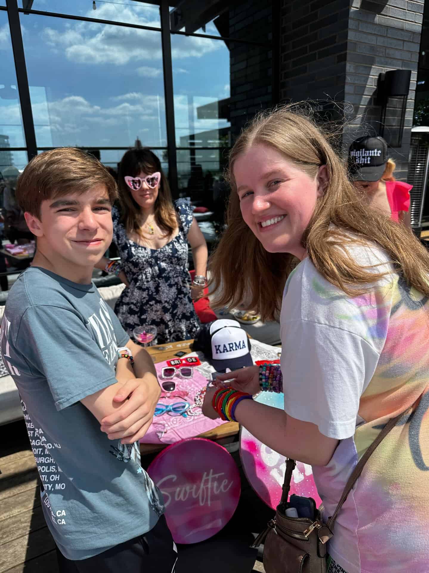 Two smiling individuals stand at an outdoor table in vibrant Nashville, showcasing colorful merchandise, including a hat labeled "Karma." A person wearing sunglasses can be seen in the background.
