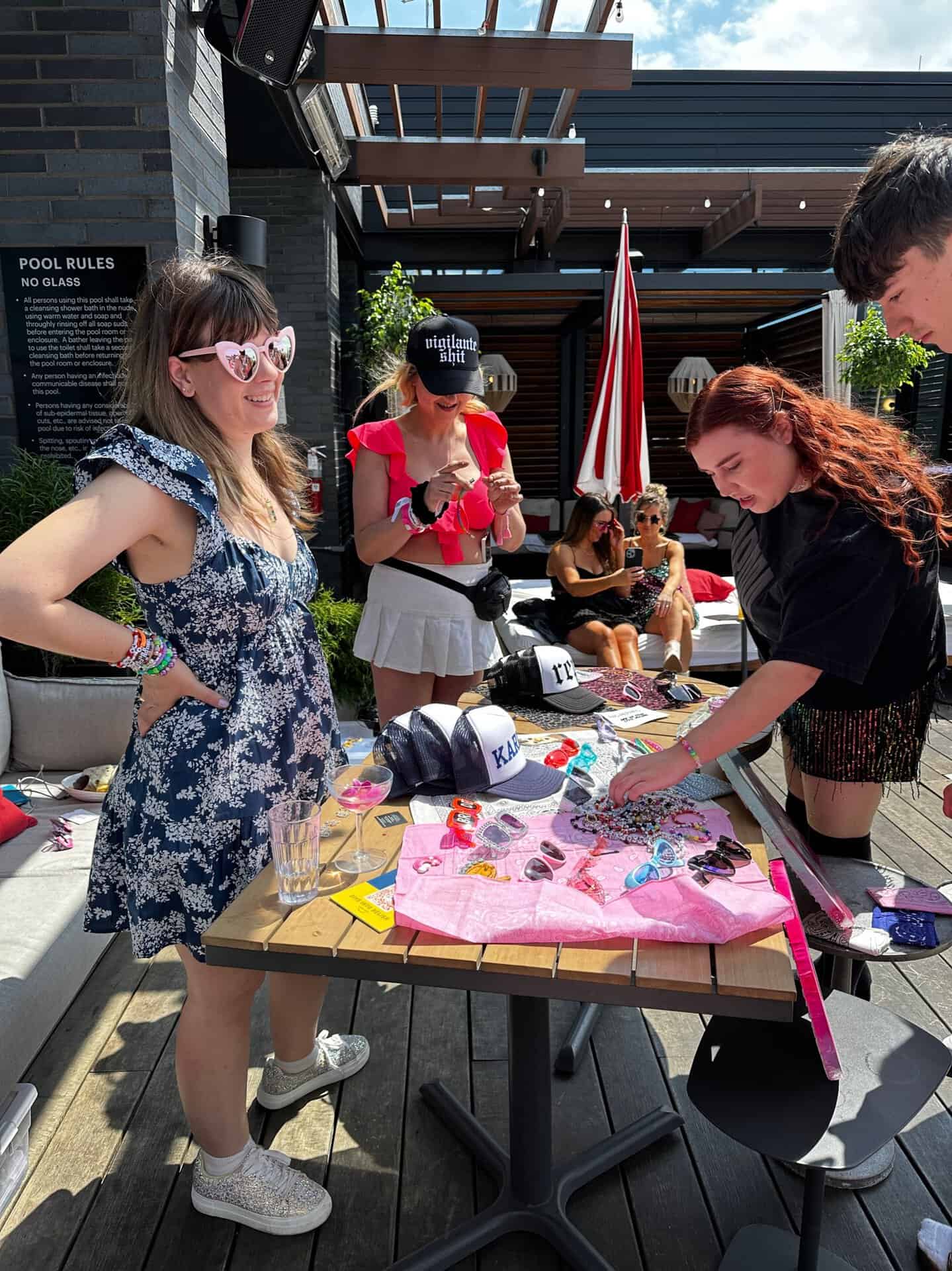 Four people gather around a table on an outdoor patio in Nashville, adorned with colorful patches and hats. A woman in a blue dress and heart-shaped sunglasses beams as her companions examine the vibrant items.