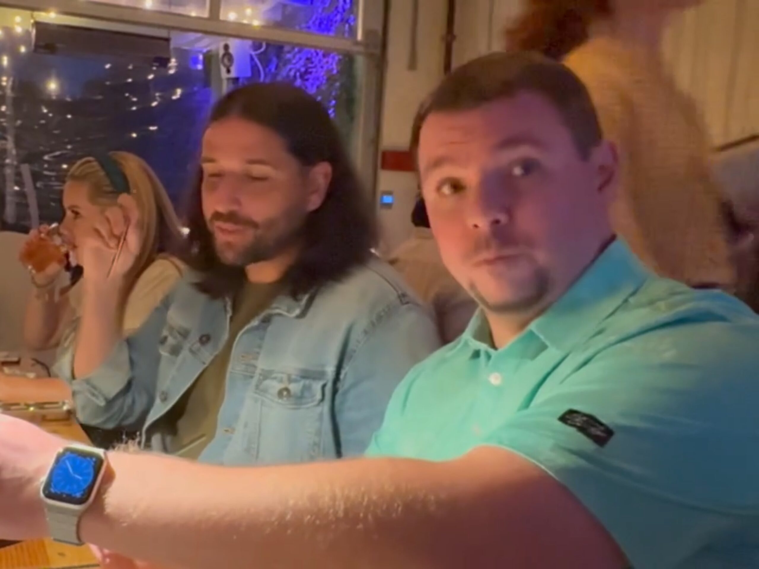 Three people sit at a restaurant table; one man in a turquoise shirt looks toward the camera while the others are engaged in conversation and drinking. String lights hang in the background.
