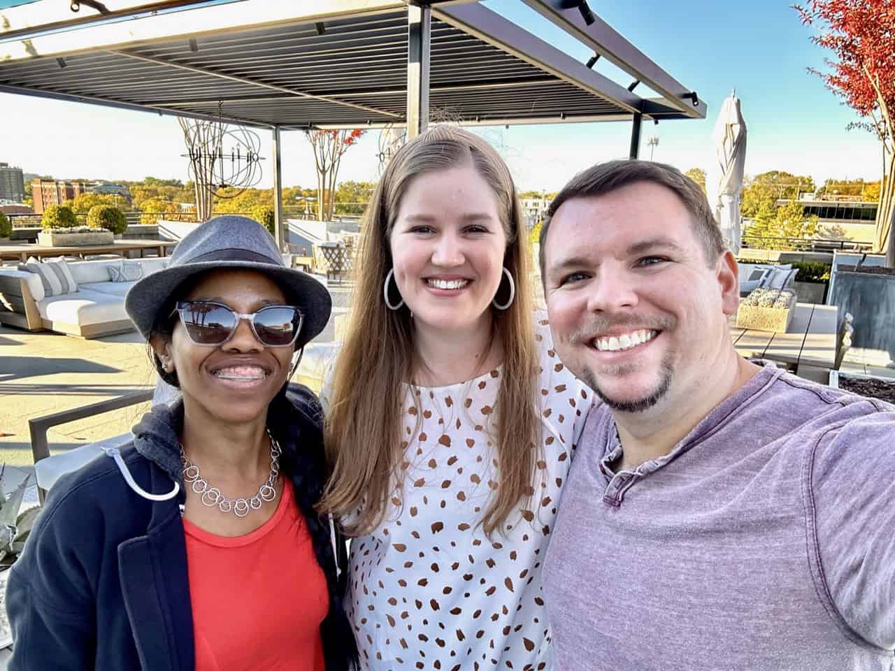 Three people smile for a selfie on a Nashville rooftop, with outdoor seating and trees adorning the skyline in the background.