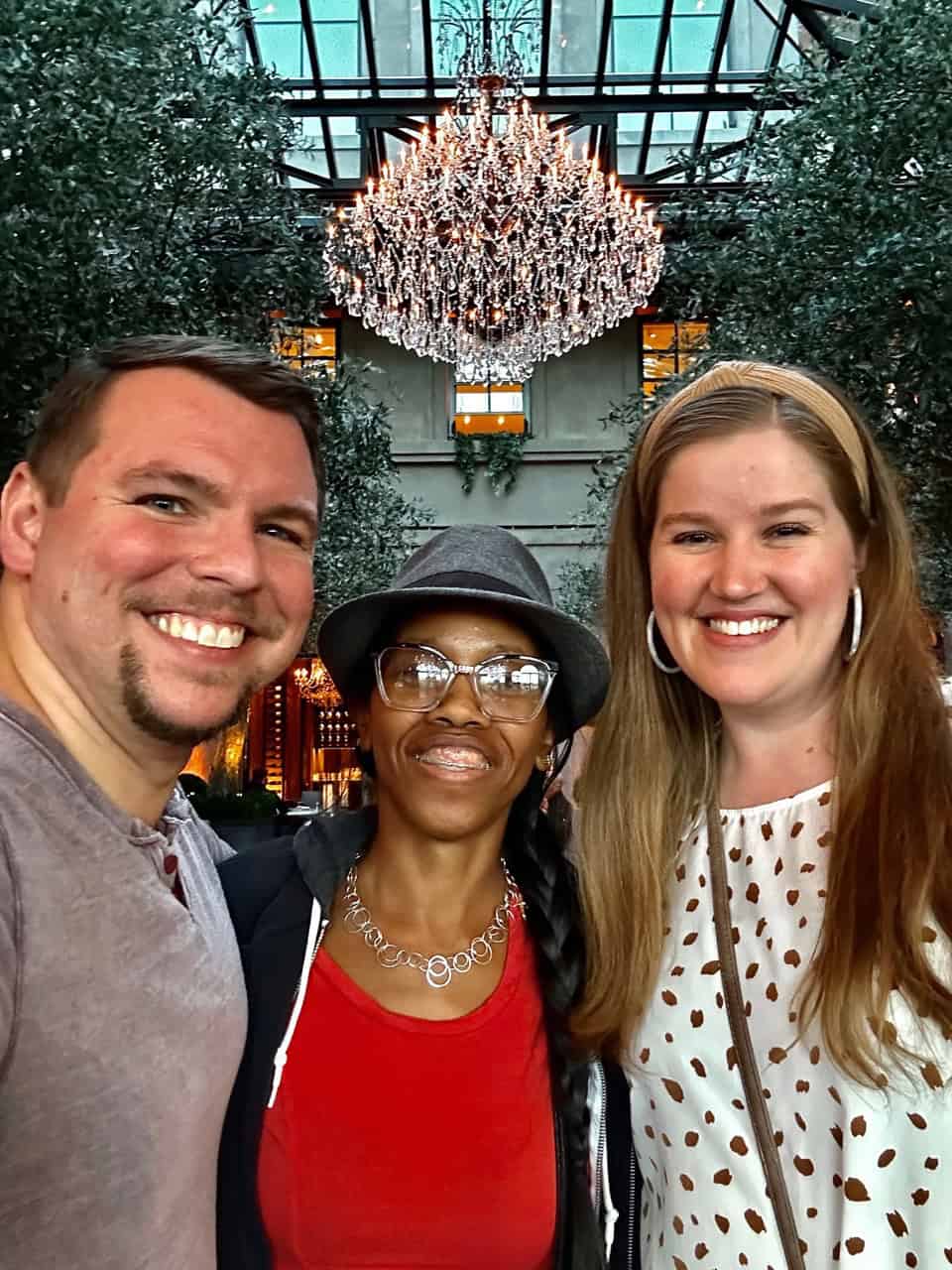 Three people smiling in a group selfie inside Nashville's iconic glass-roofed hall, with a dazzling chandelier hanging above.