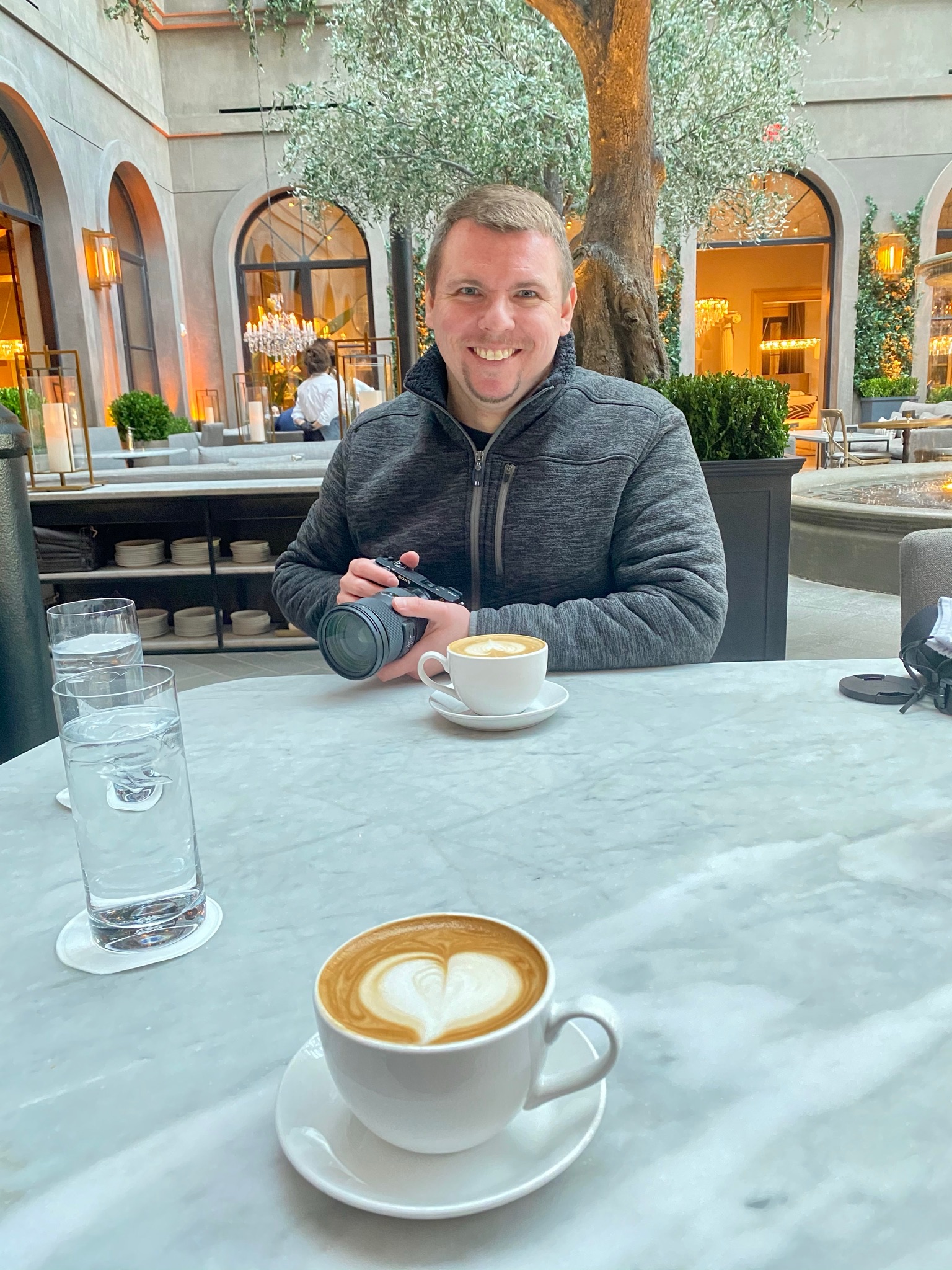 A person sits at an outdoor café table in Nashville, holding a camera, with two cups of coffee and a glass of water on the table. The courtyard setting features lush greenery and arched windows.