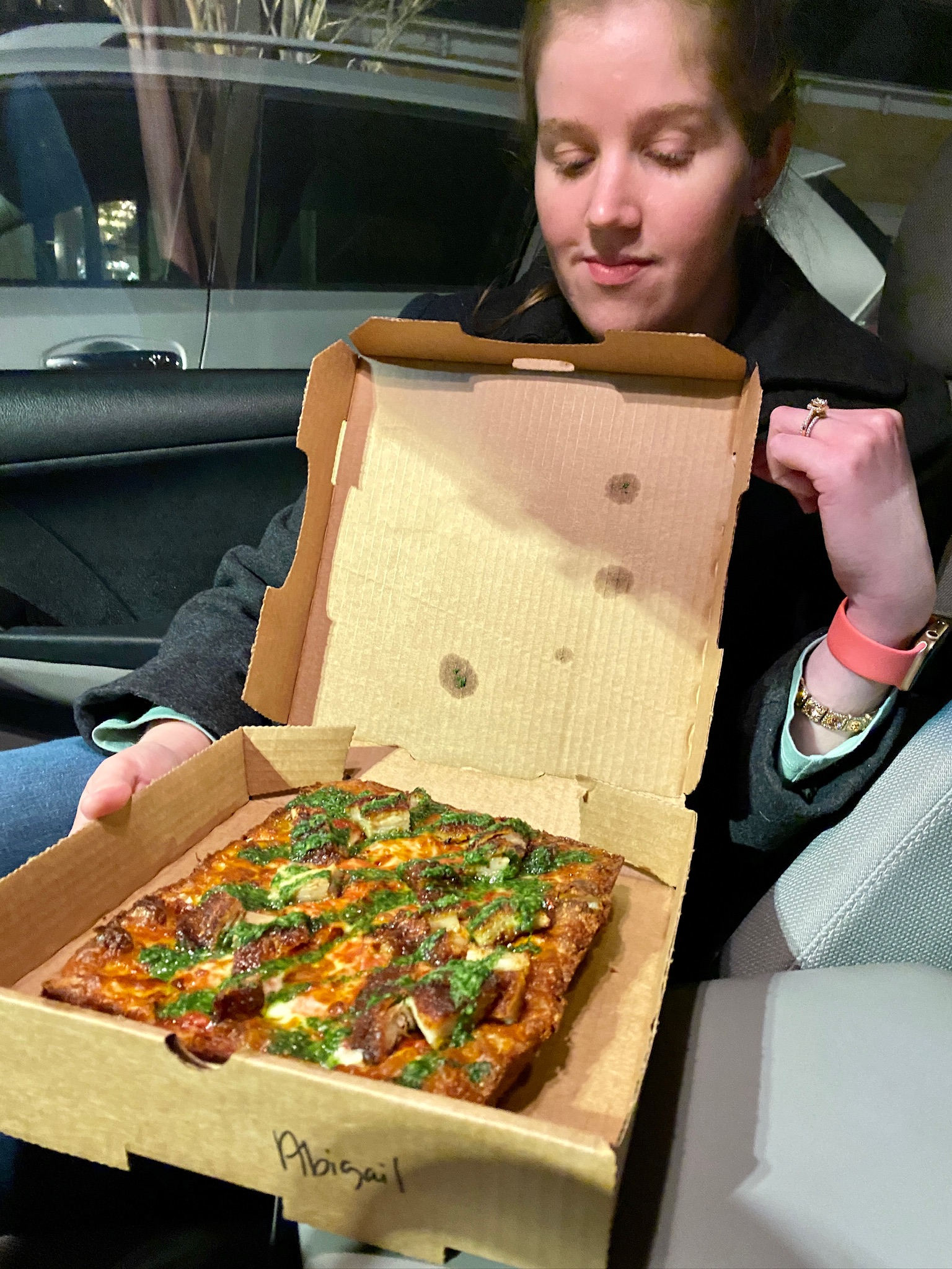 A person sitting in a car holds an open pizza box with a rectangular Nashville-style pizza topped with green herbs and chunks of meat or vegetables.