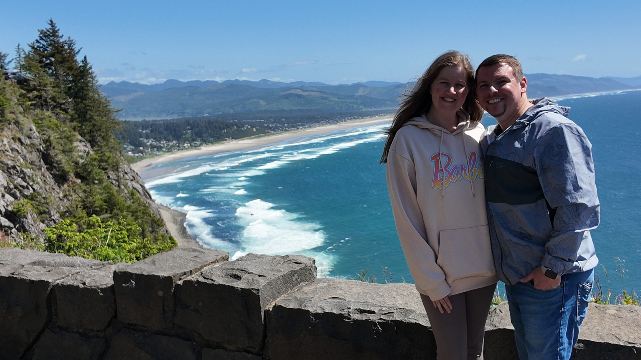 A smiling couple poses in front of a stone wall with an ocean view, waves, and a coastline with distant hills and a partly cloudy sky in the background.