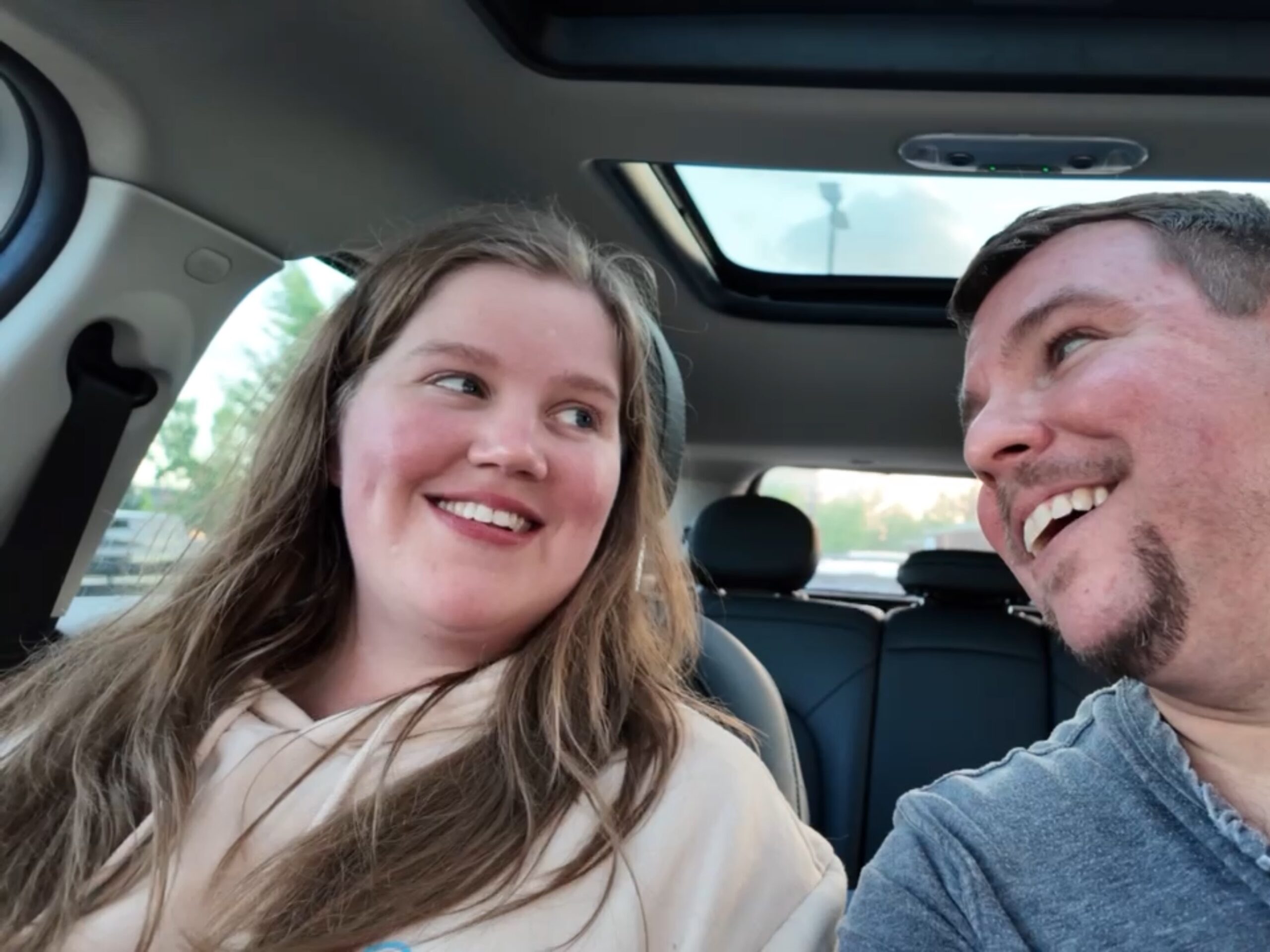 Two people are sitting in a car, looking at each other and smiling. The car has a sunroof, and it appears to be daytime outside.