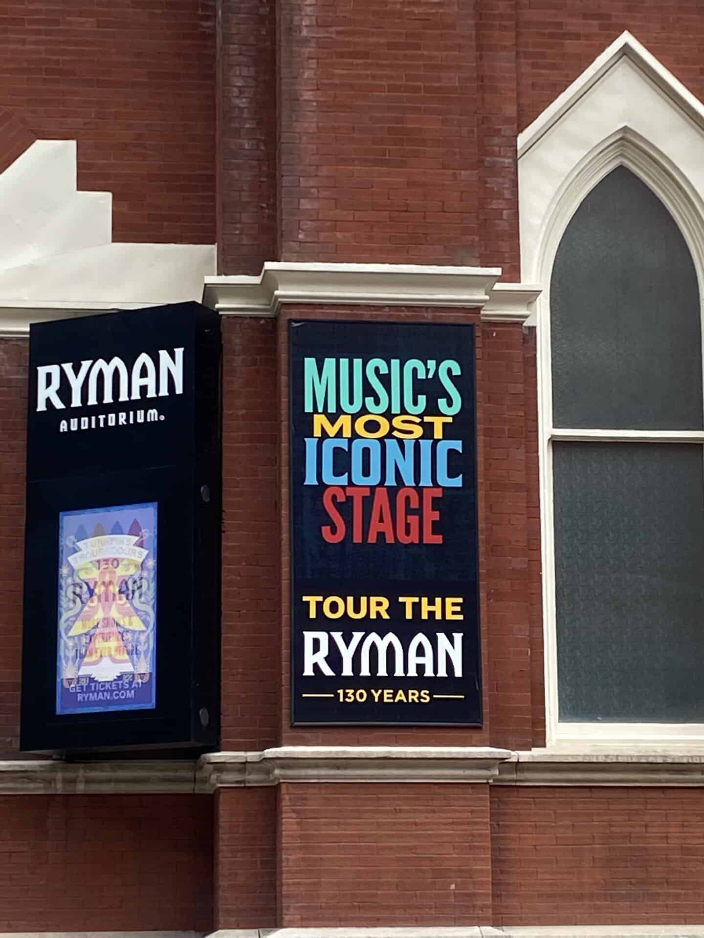 Sign on a brick building reads, "Music's Most Iconic Stage. Tour the Ryman. 130 Years," next to a Ryman Auditorium poster.