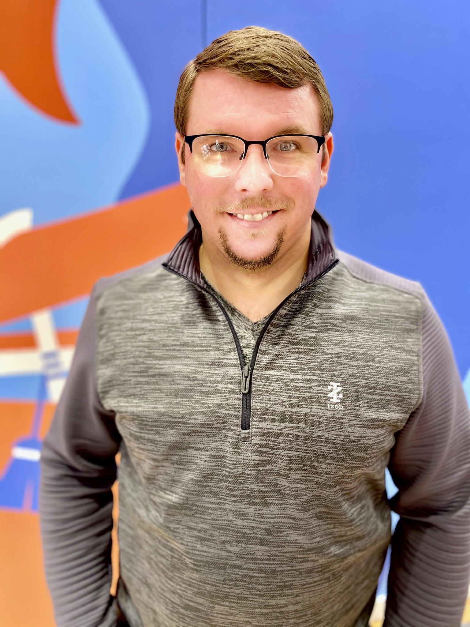A man with short brown hair and glasses wearing a gray quarter-zip pullover stands in front of a blue and orange abstract background, smiling at the camera.