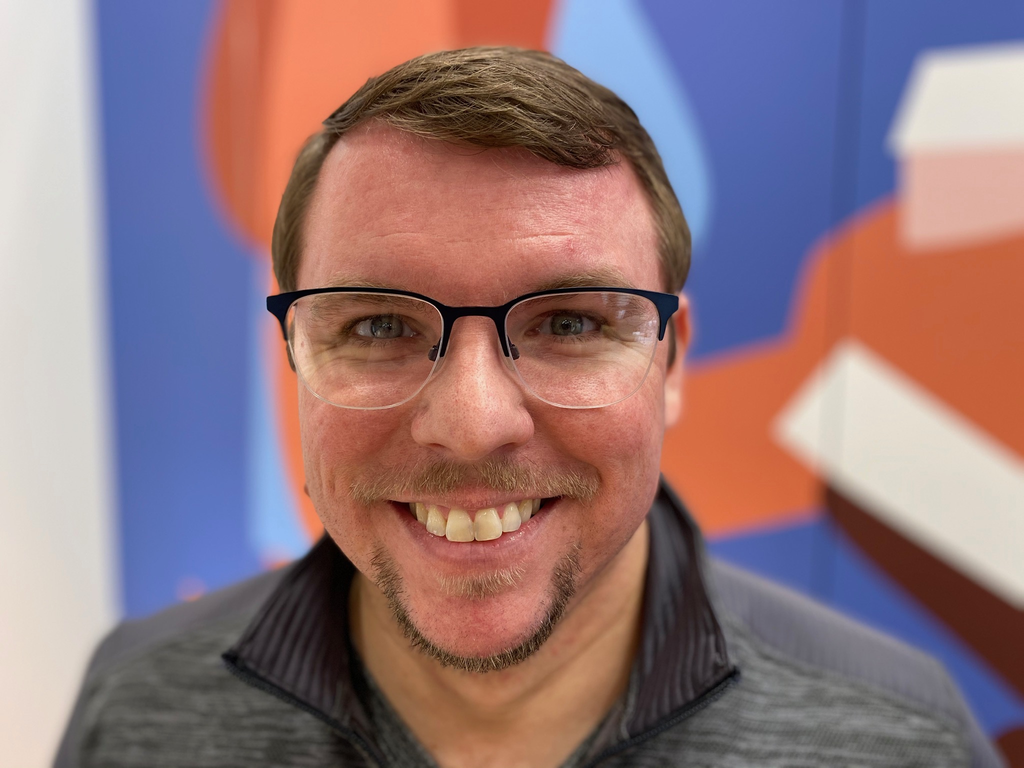 A man wearing glasses and a gray shirt smiles at the camera, standing in front of a colorful abstract background.