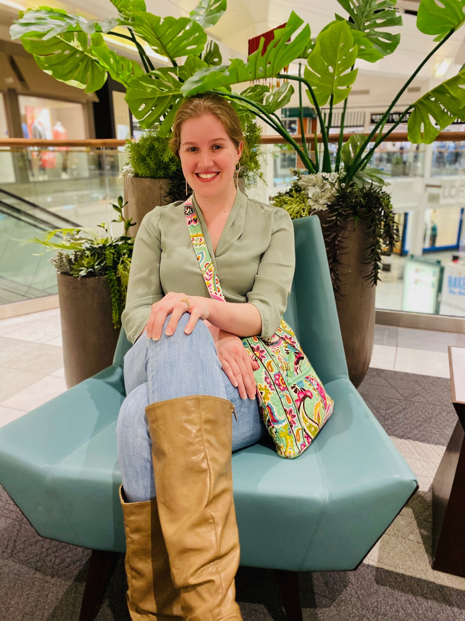 A woman with light brown hair sits on a light blue chair in a mall, wearing a green blouse, jeans, tan boots, and holding a colorful patterned bag. Large potted plants are behind her.