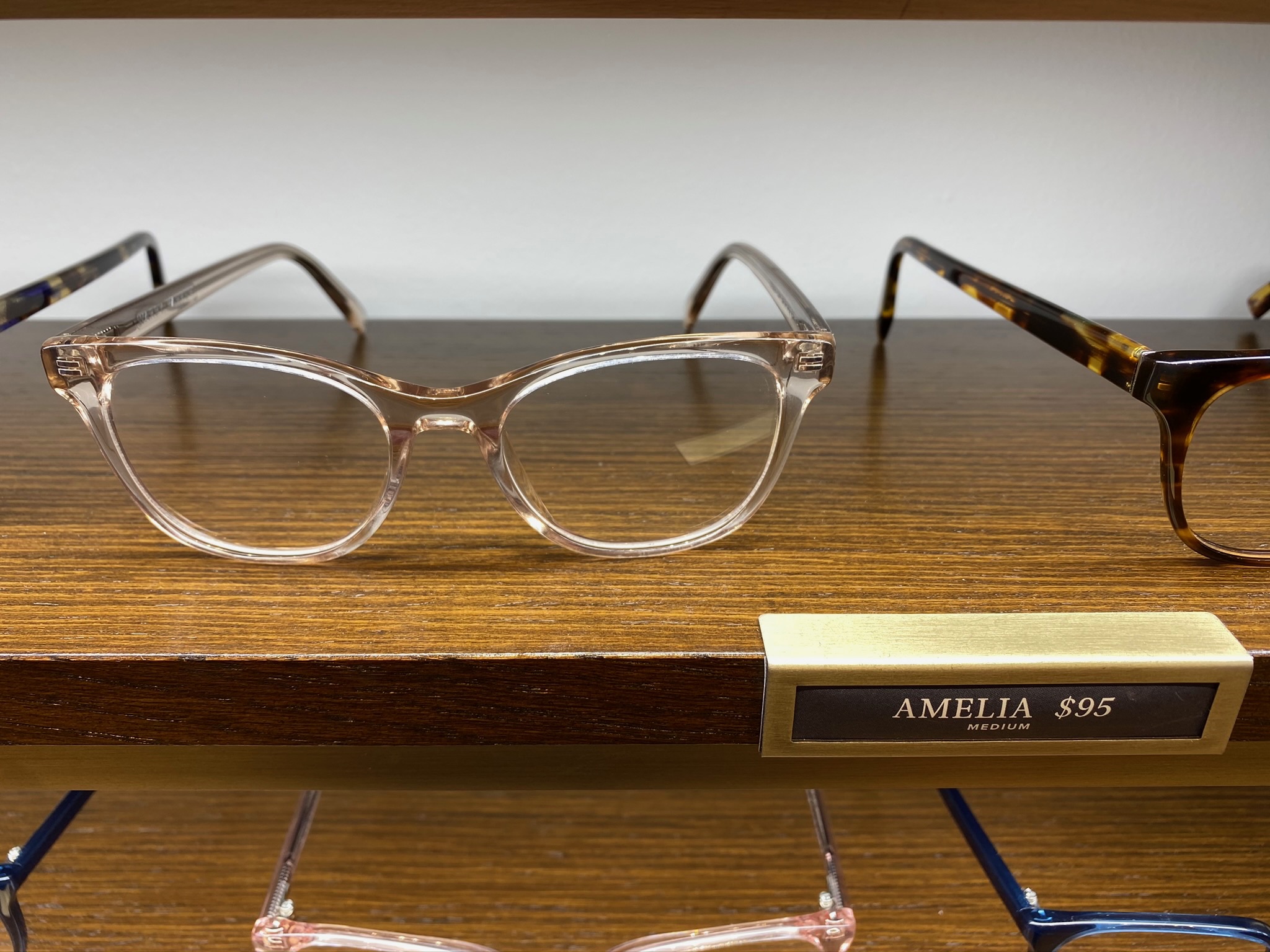 A pair of clear, square-framed eyeglasses labeled "AMELIA $95 MEDIUM" displayed on a wooden shelf.
