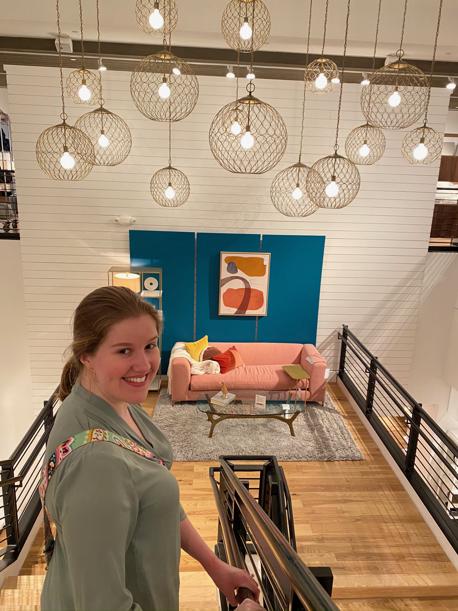 A woman stands on a staircase overlooking a modern living room with a pink sofa, abstract art, and hanging globe lights.