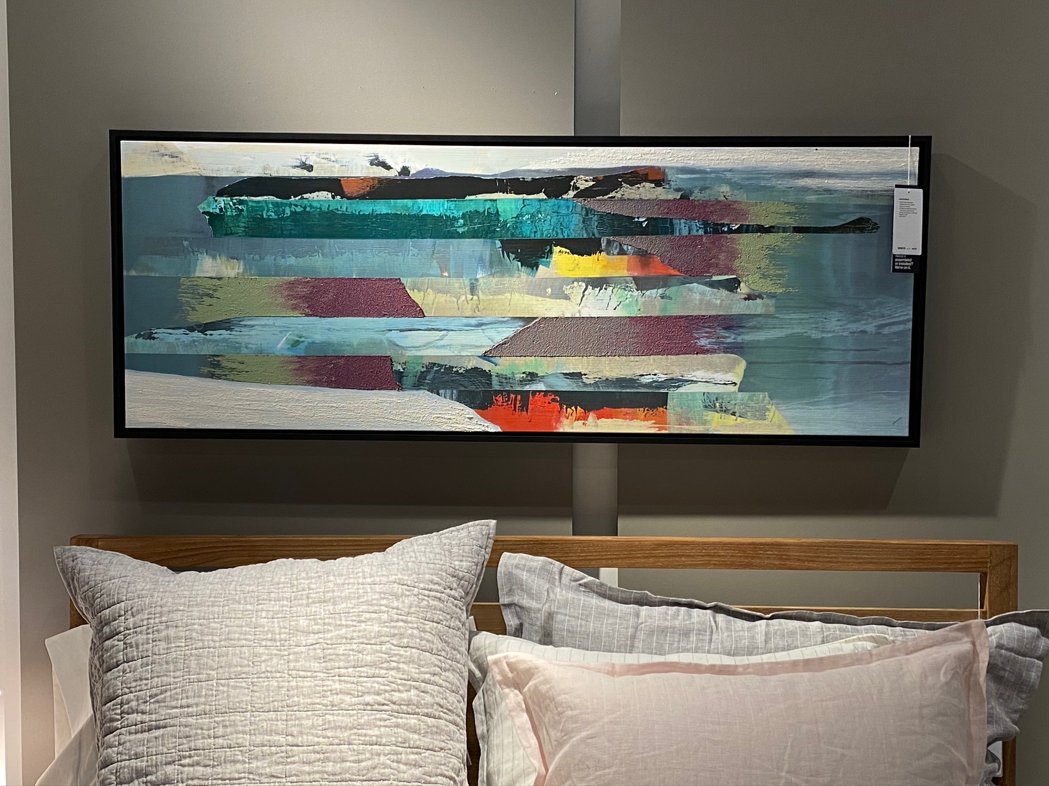 A rectangular abstract painting with layered horizontal brushstrokes in various colors hangs above a bed with gray and blush pillows.