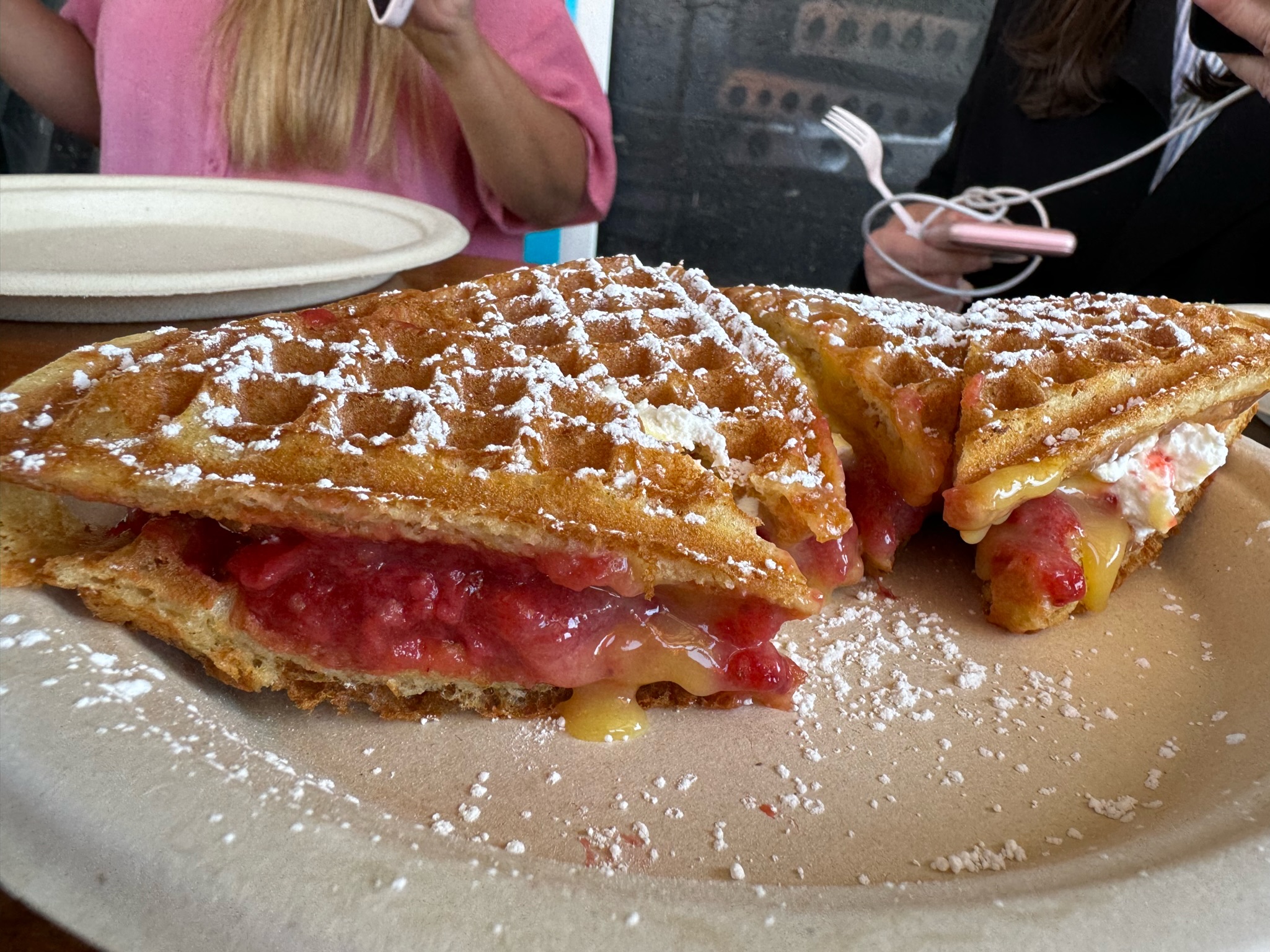 Two triangular waffles dusted with powdered sugar, filled with fruit and cream, are served on a paper plate; two people sit in the background holding utensils.