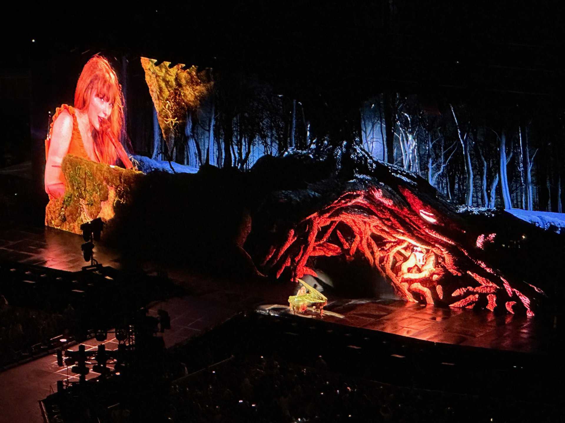 A Nashville performer graces the piano on stage, a large screen behind displaying a serene forest scene with illuminated red tree roots.