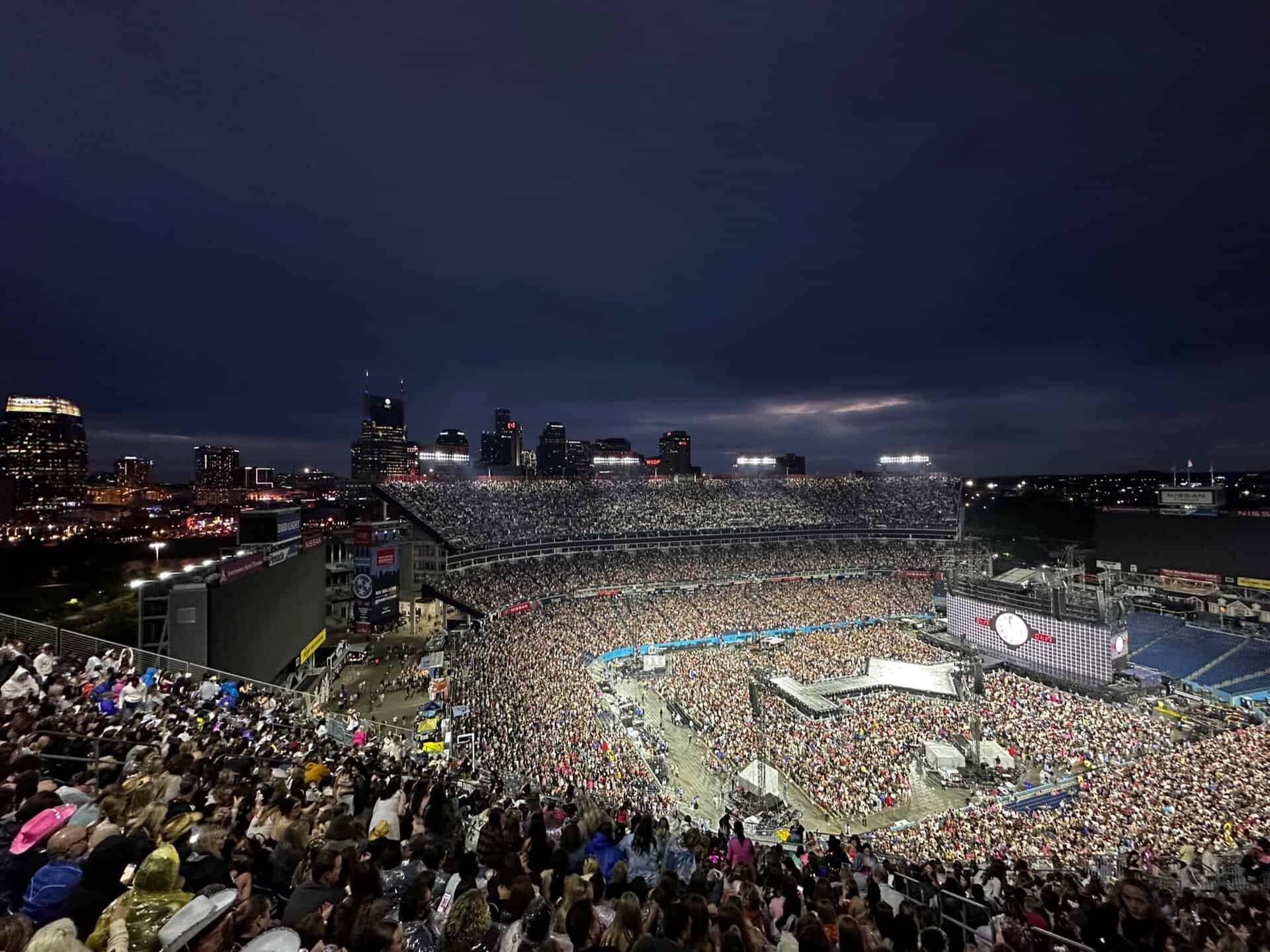 A large crowd fills the Nashville stadium at night, with the illuminated city skyline providing a stunning backdrop. Stage and screens are set up for a concert, creating an electric atmosphere in Music City.