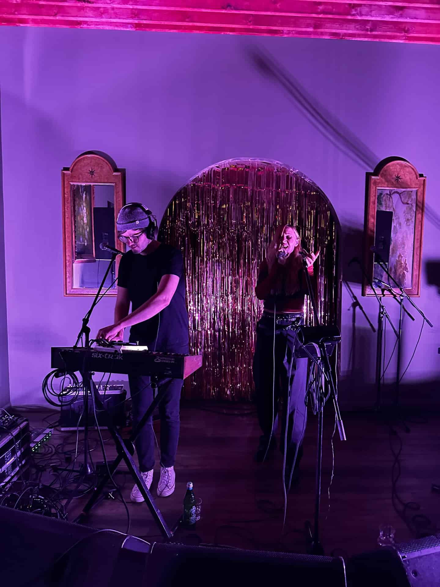 Two musicians perform on stage with a keyboard and microphones, enveloped in Nashville's vibrant energy, surrounded by purple lighting and a gold tinsel backdrop.