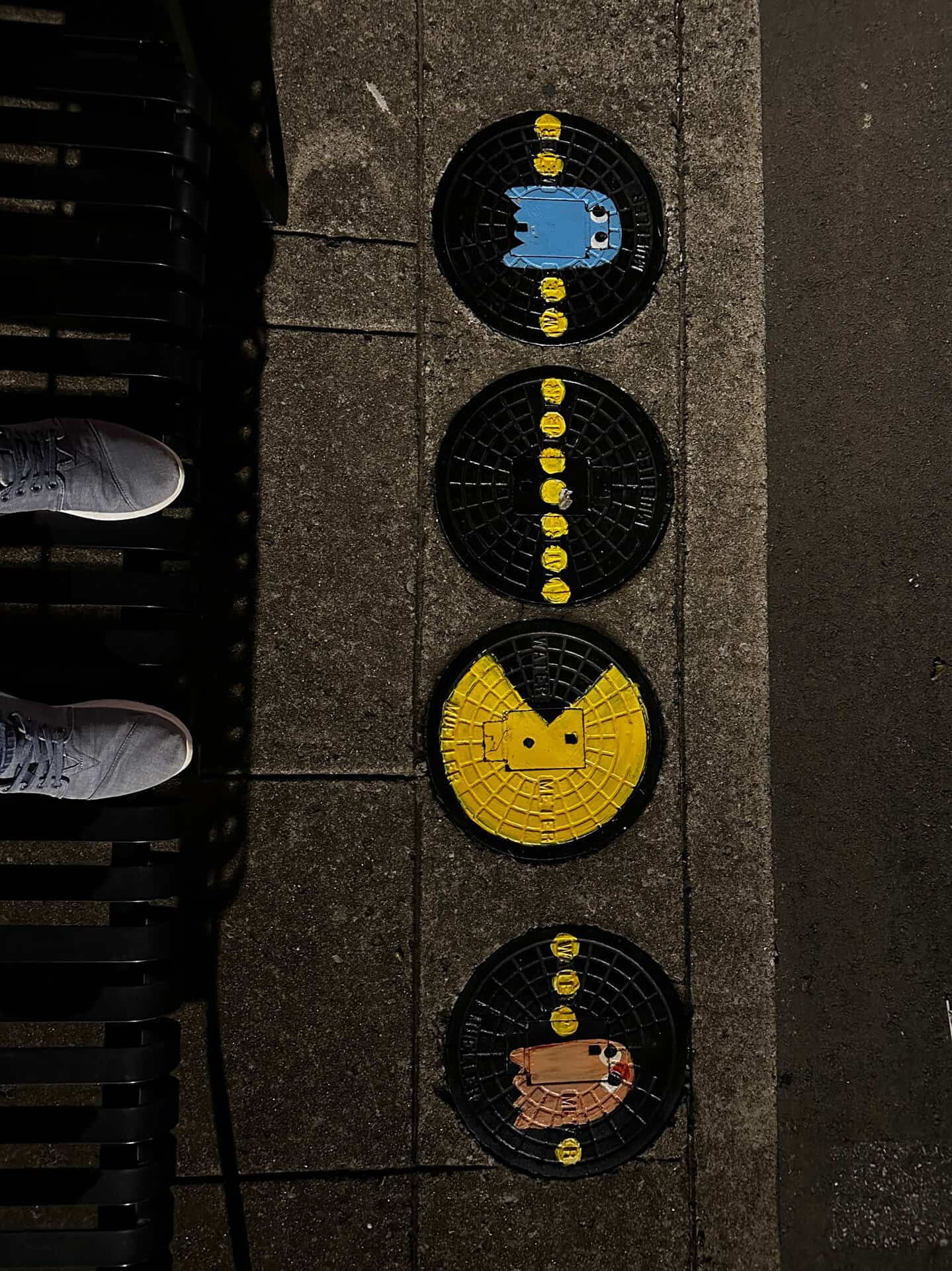 Four sewer covers in Nashville painted with Pac-Man game elements, showcasing Pac-Man, a blue ghost, and dots. A person's feet in sneakers can be seen to the left.