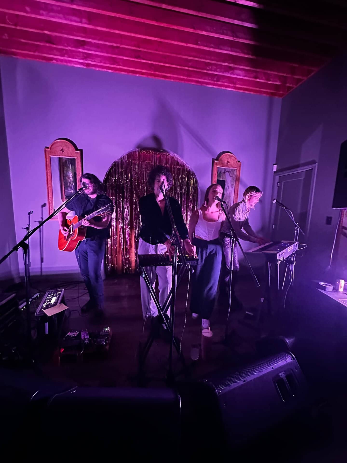 Four Nashville musicians perform on stage with purple lighting, featuring guitarists and keyboard players. A decorative backdrop with gold fringe and arched wall panels completes the vibrant scene.