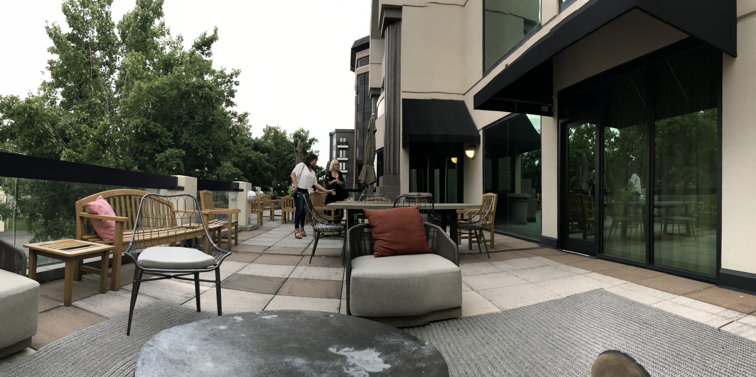 A modern outdoor patio with wooden furniture, a round table, cushioned chairs, and several people standing and talking near a building with large glass windows.