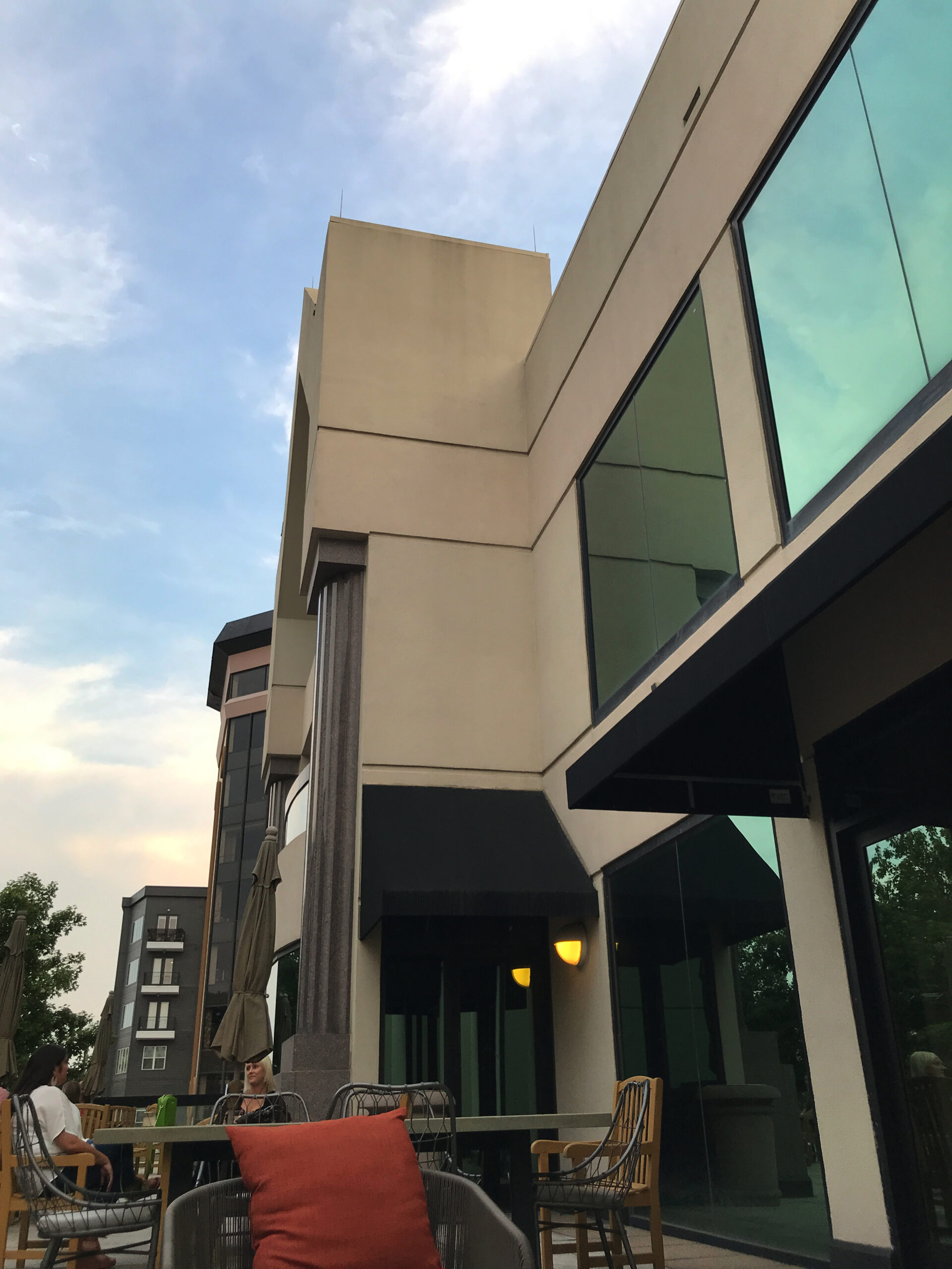 Outdoor seating area with chairs and tables next to a modern building with large windows and awnings, photographed in the early evening.