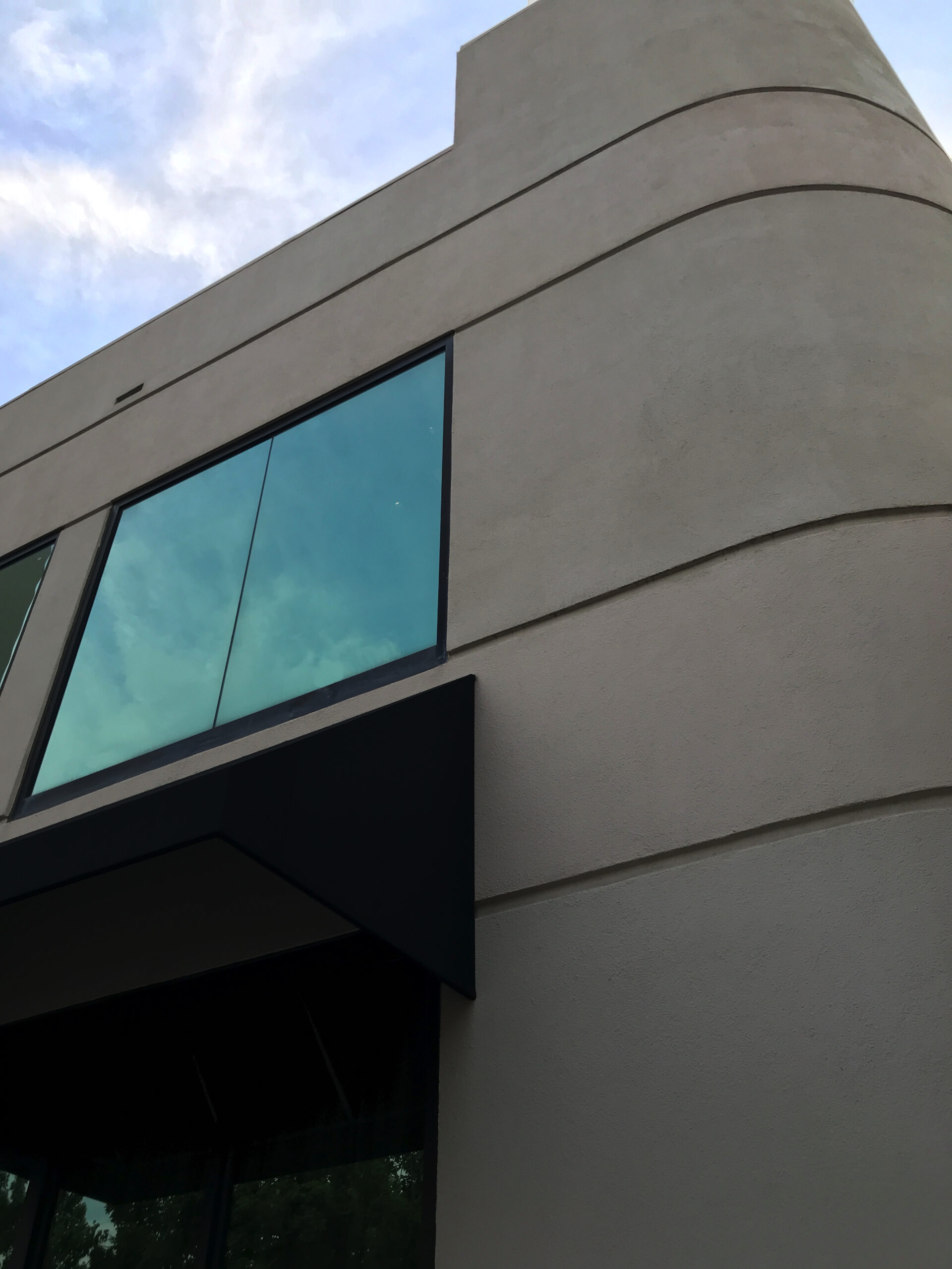 A modern beige building with large glass windows and a black awning, viewed from a low angle against a partly cloudy sky.