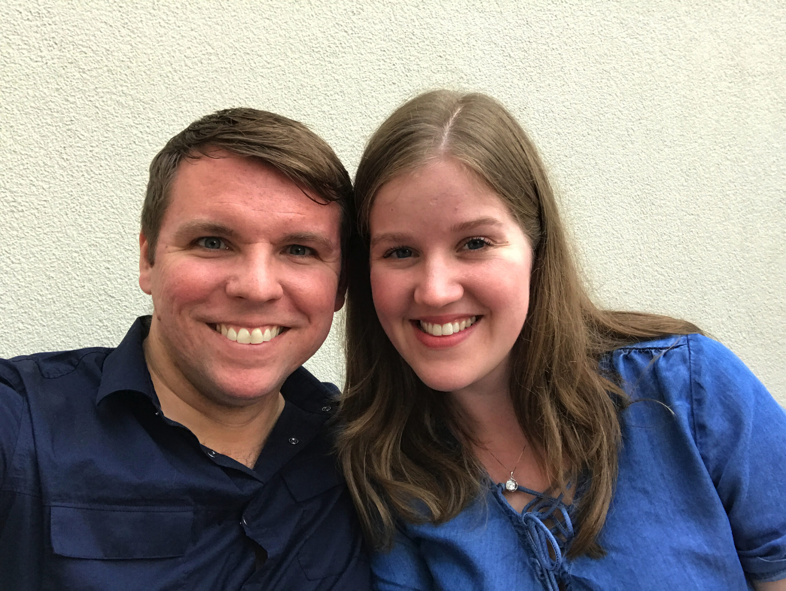 A man and a woman smiling at the camera, both wearing blue tops, standing in front of a plain light-colored wall.