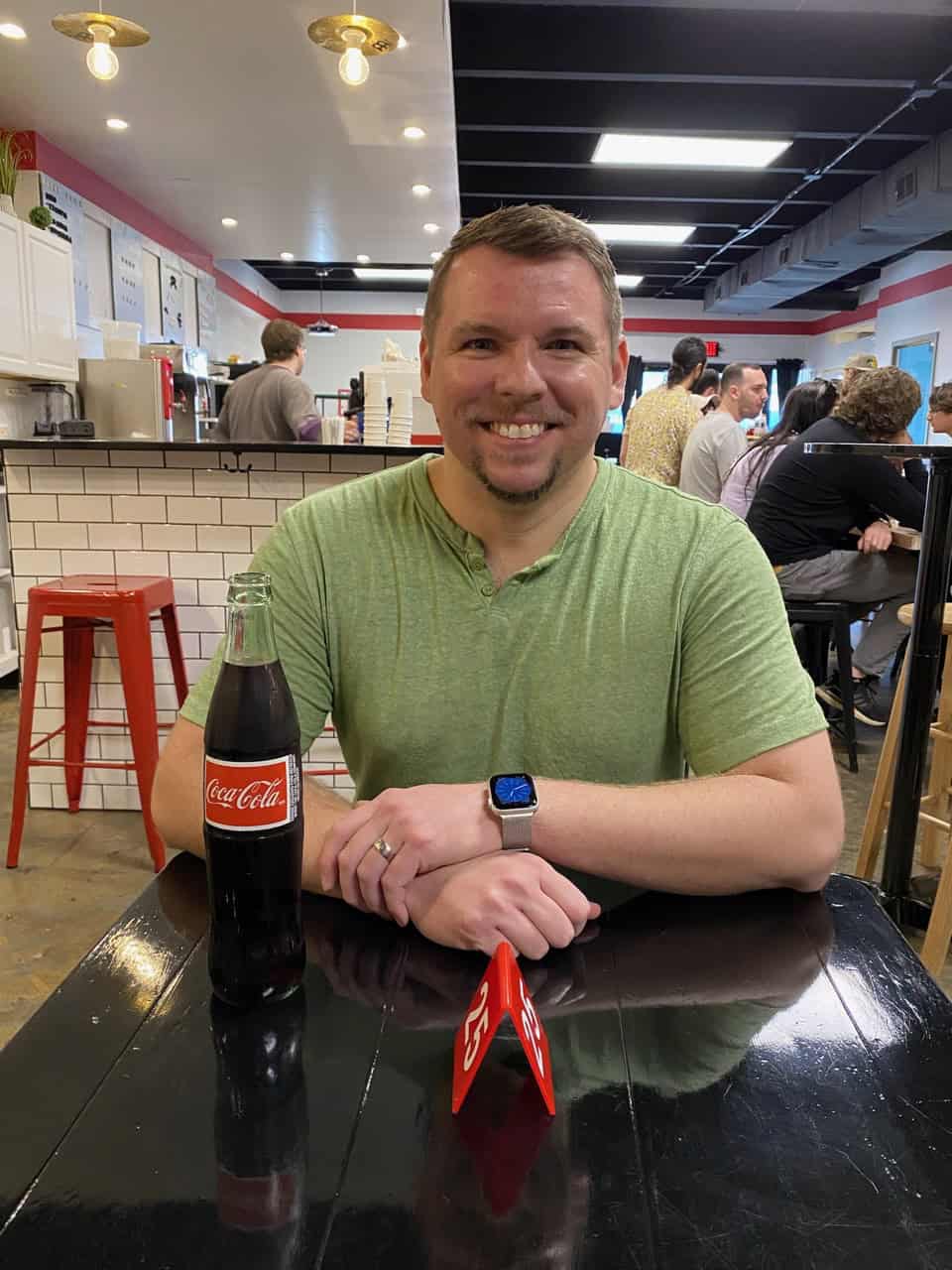 A person in a green shirt sits at a table with a bottle of Coca-Cola and a red card, evoking the lively vibe of Nashville. Other people and a black chair are visible in the background.