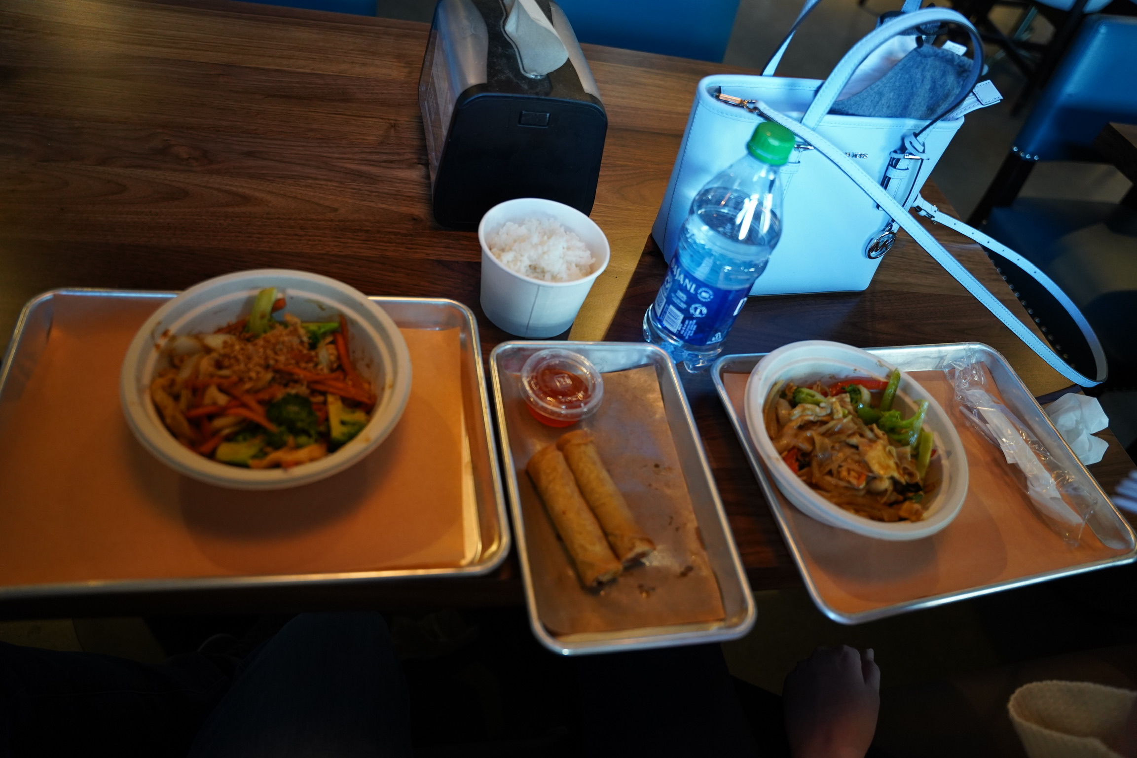 Two trays with bowls of stir-fried noodles and vegetables, two spring rolls with sauce, a cup of rice, a bottle of water, and a white handbag on a wooden table create the perfect Nashville lunch spread.
