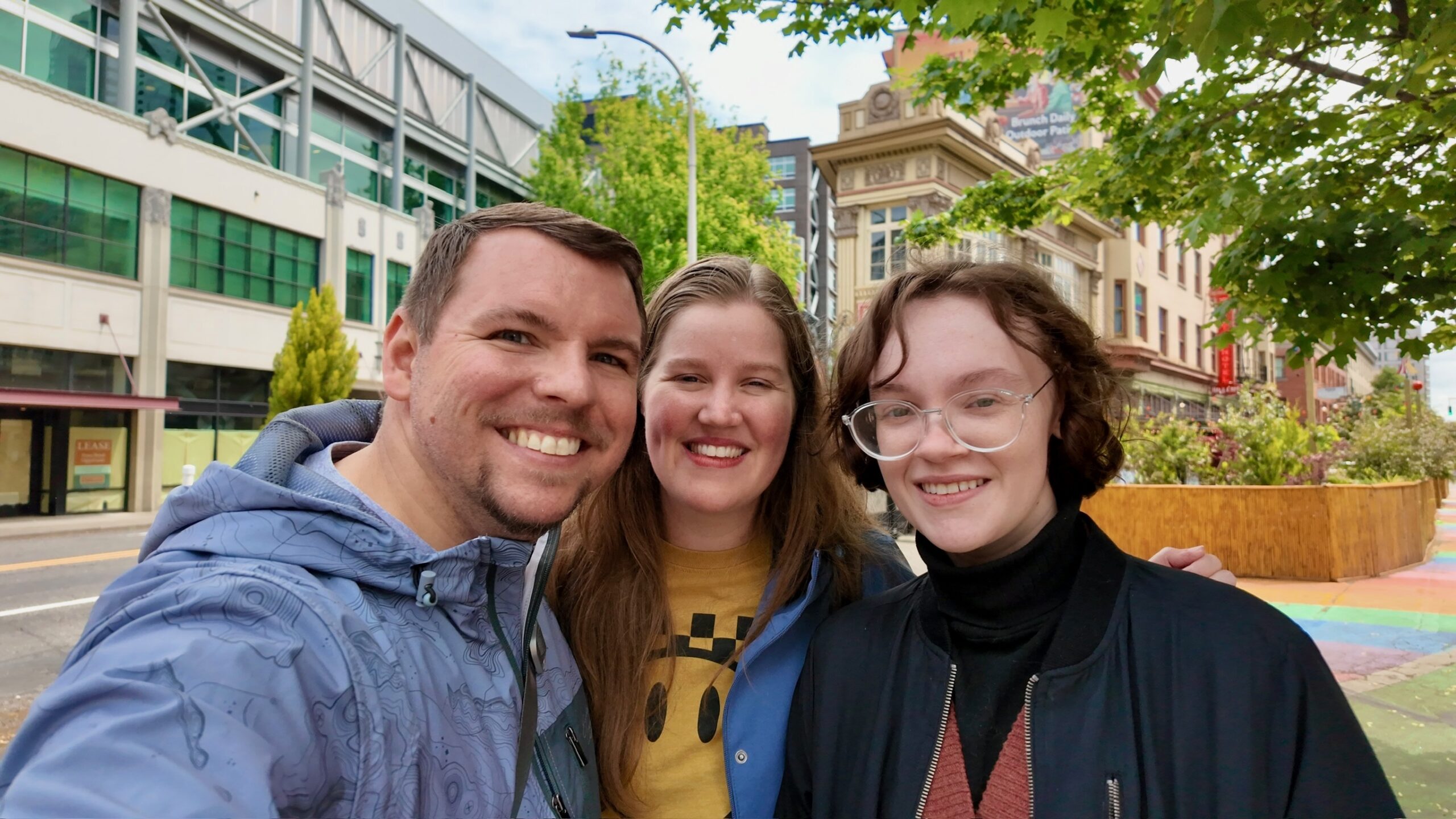 Three people are smiling while taking a selfie together outdoors on a city street with buildings and trees in the background.