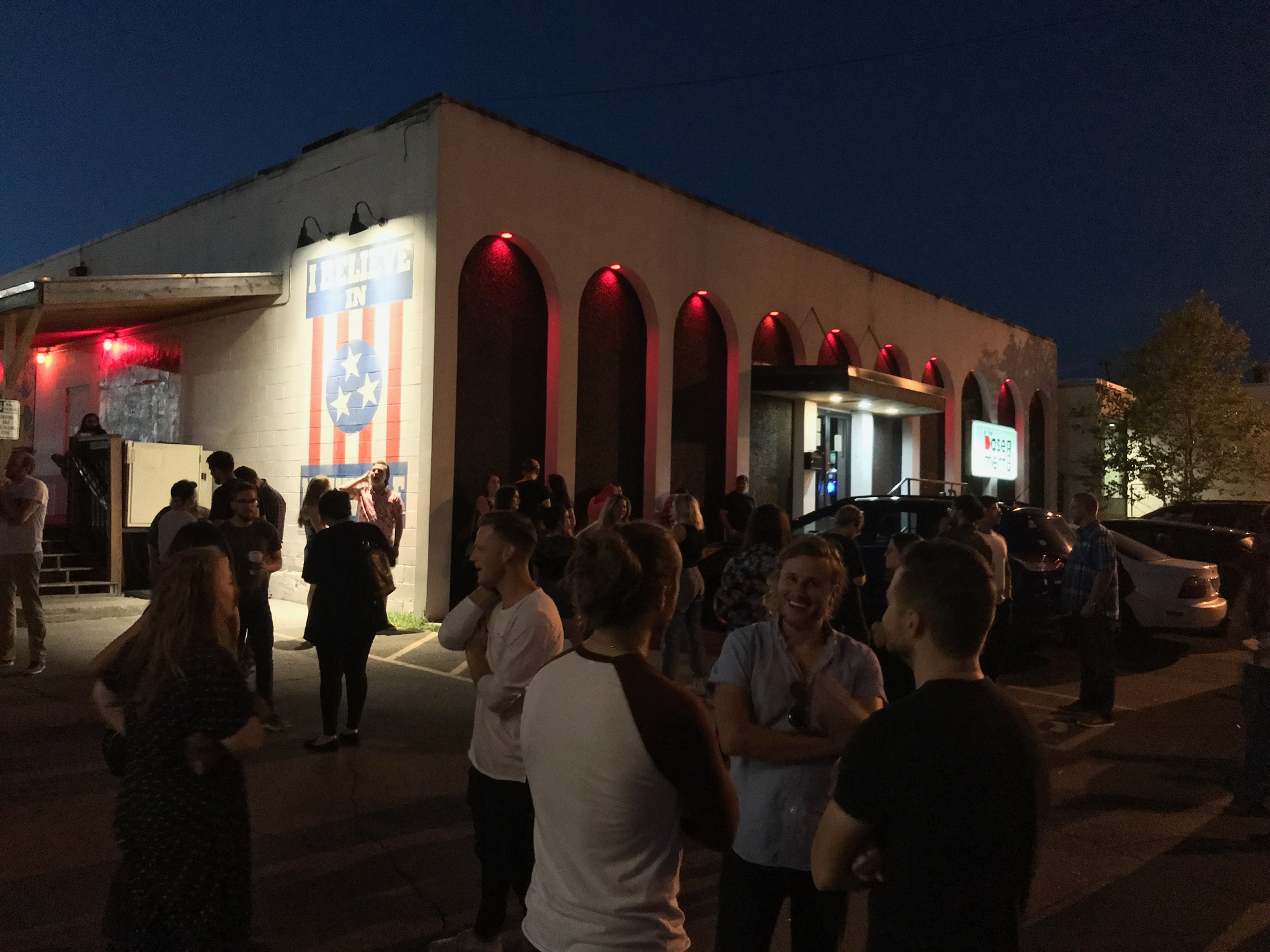 A group of people gathers outside a building at night under red lights, with a Tennessee flag mural and parked cars visible.