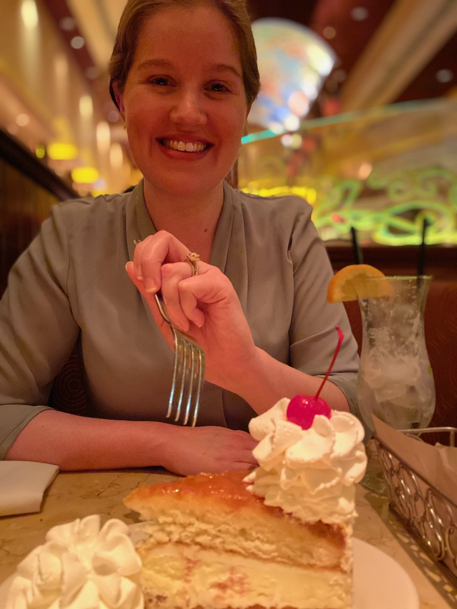 A woman smiles at a restaurant table, holding a fork above a slice of cake topped with whipped cream and a cherry; a drink with a lemon slice is beside her.
