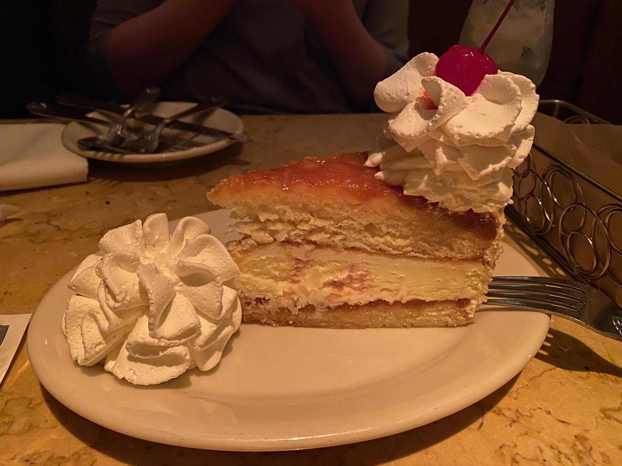 A slice of layered cake with whipped cream and a cherry on top, served on a plate with an extra dollop of whipped cream and a fork.