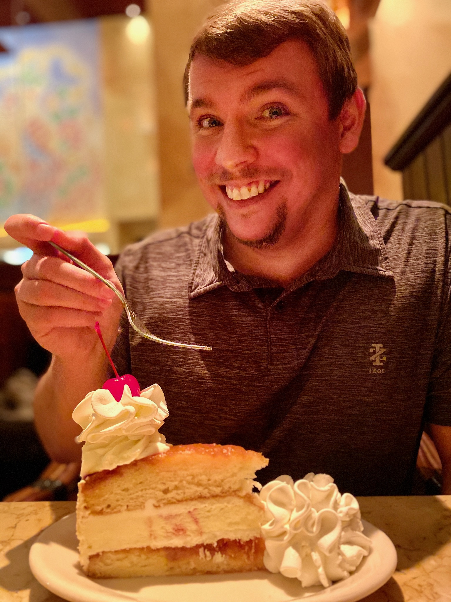A man smiling and holding a fork above a slice of layered cake with whipped cream and a cherry at a restaurant table.