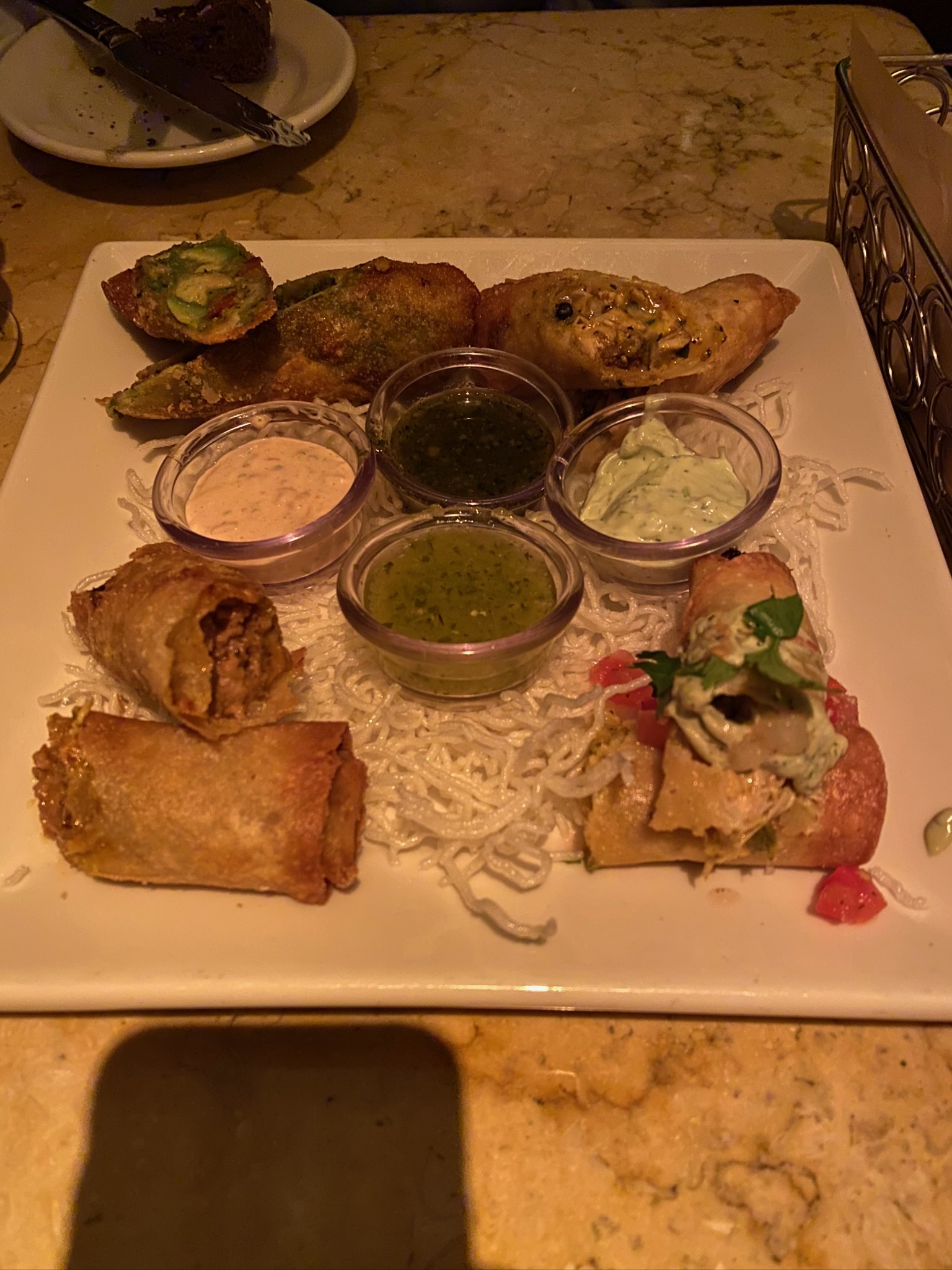 A square plate with assorted appetizers, including spring rolls and stuffed items, served with three different dipping sauces in small cups, on a bed of crispy noodles.