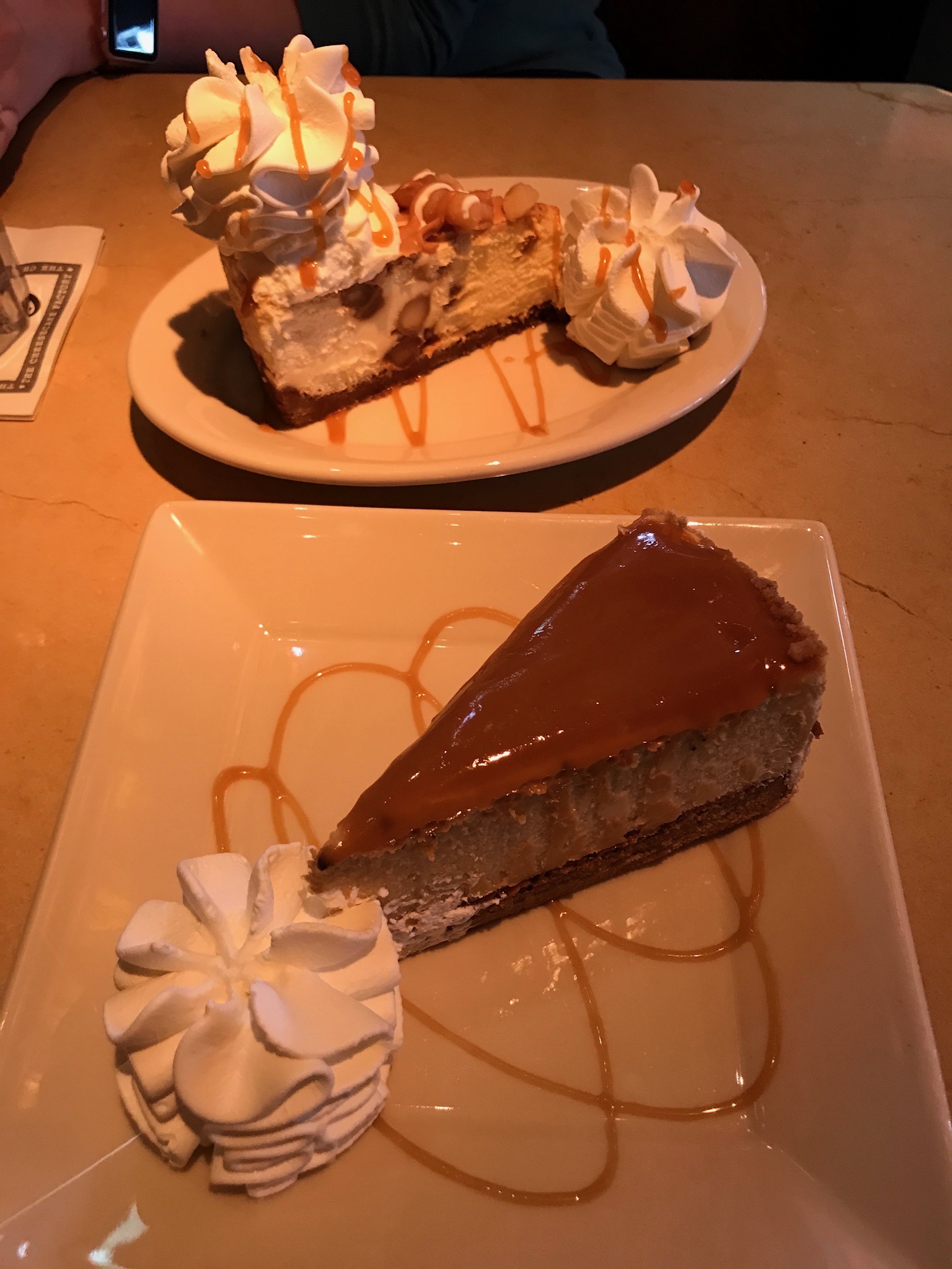 Two slices of cheesecake on plates, each topped with caramel sauce and served with whipped cream. One slice is in the foreground, while the other is in the background.