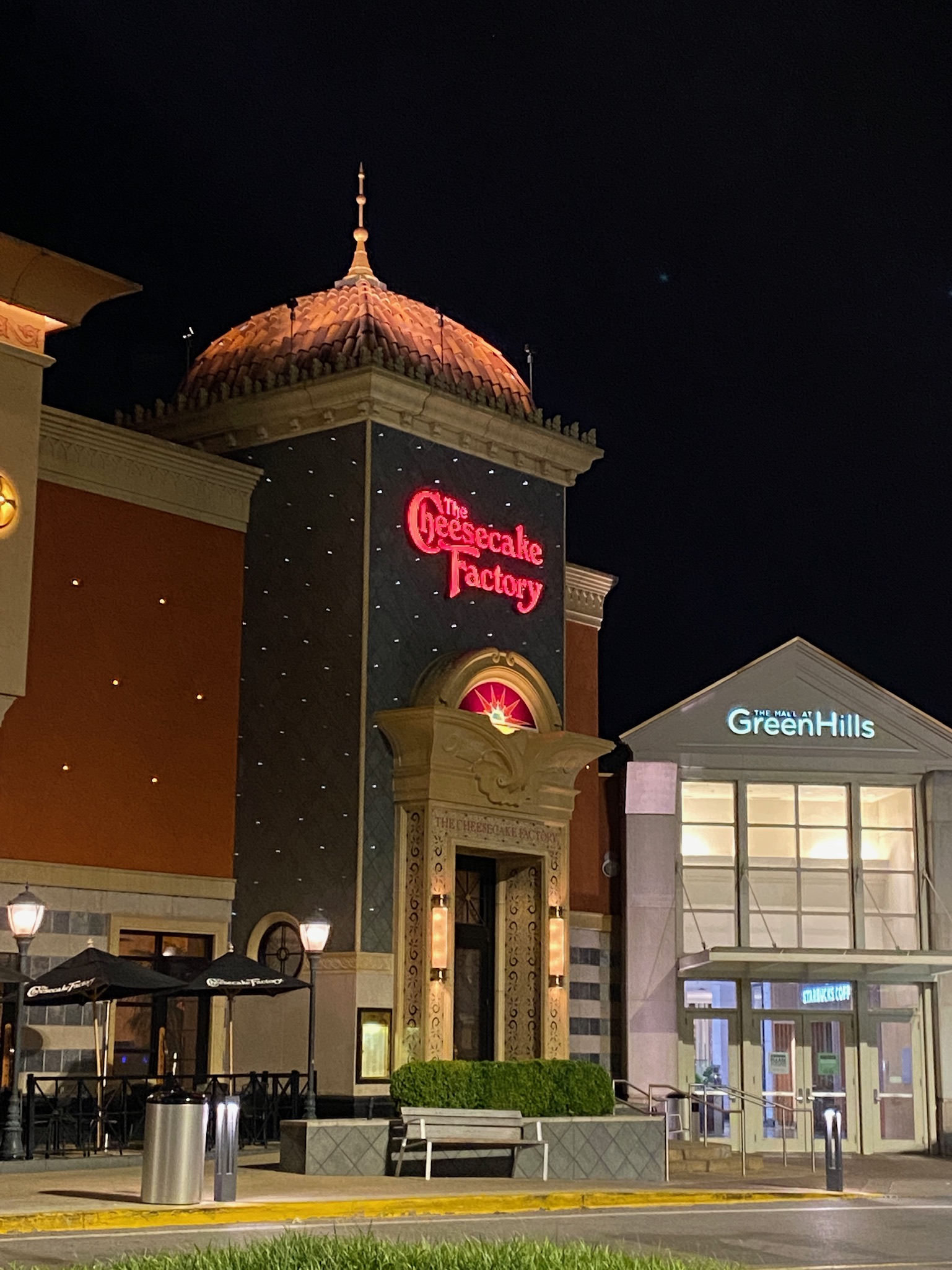 The Cheesecake Factory restaurant entrance is lit up at night next to the mall entrance for The Mall at Green Hills.