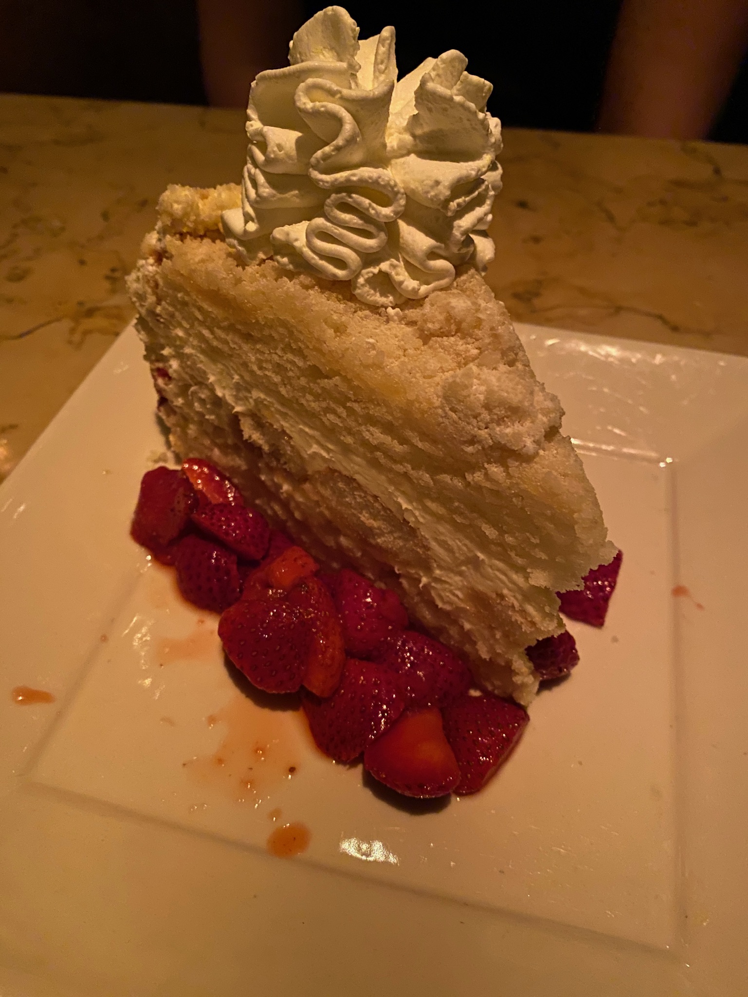A large slice of white layered cake with whipped cream on top, served with sliced strawberries on a white square plate.