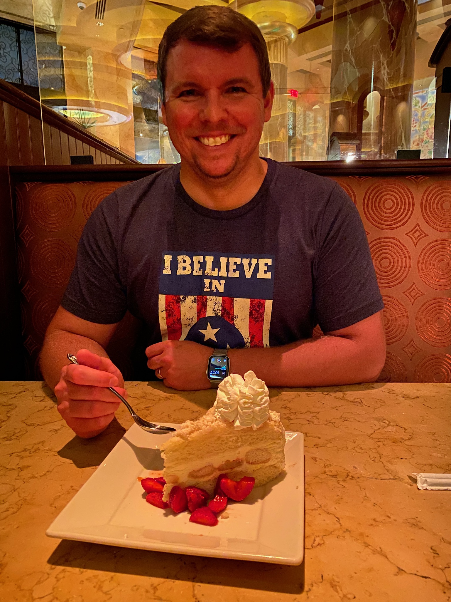 A man sits at a restaurant booth, smiling at the camera, holding a fork over a plate with cake, whipped cream, and strawberries.