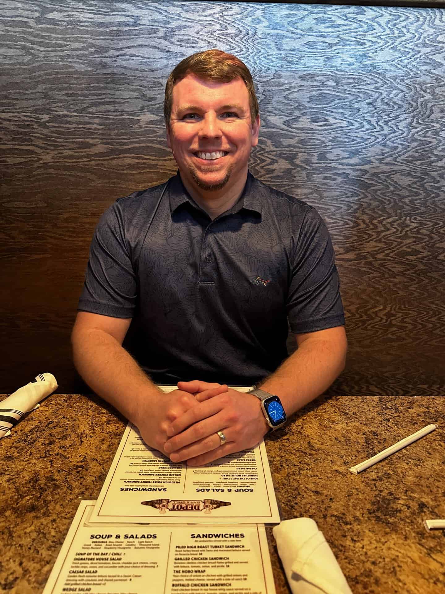 A person sitting at a Nashville restaurant table with menus, wearing a dark shirt and a smartwatch, smiling at the camera.