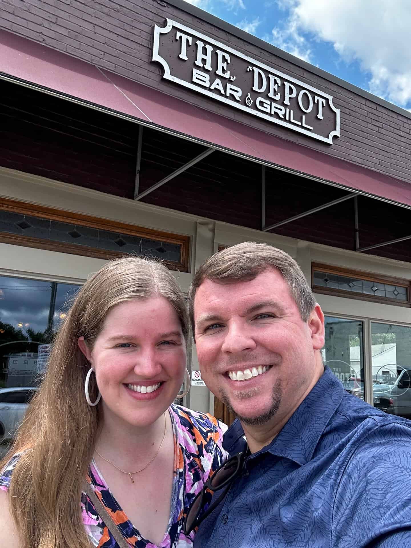 Two people smiling in front of The Depot Bar & Grill near Nashville. The man wears a blue shirt, and the woman sports a floral top. A sign is visible in the background.