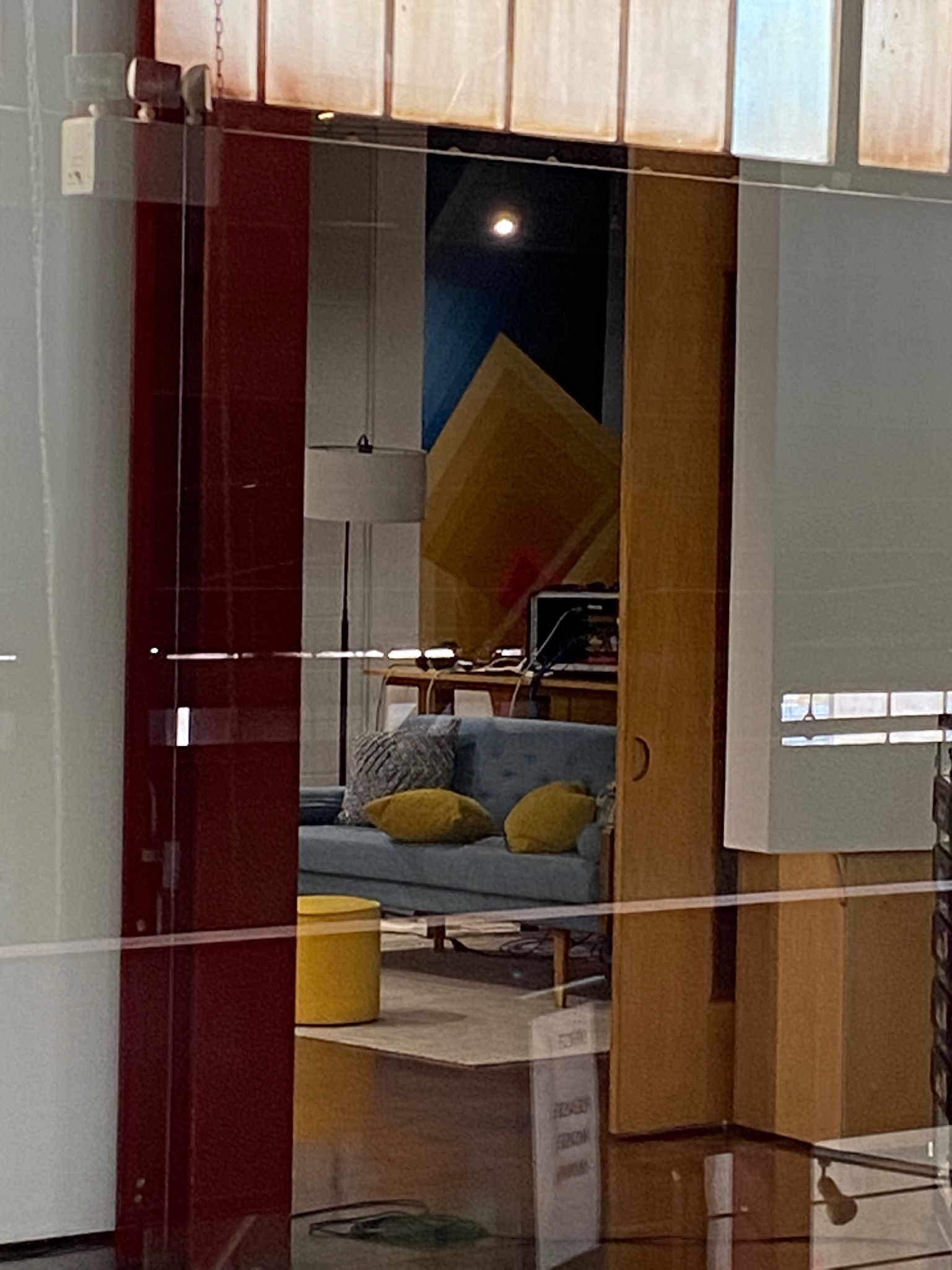 Photo of a modern office lounge with a blue sofa, yellow pillows, a yellow ottoman, a wooden table, and geometric wall art. The scene is viewed through a glass partition.