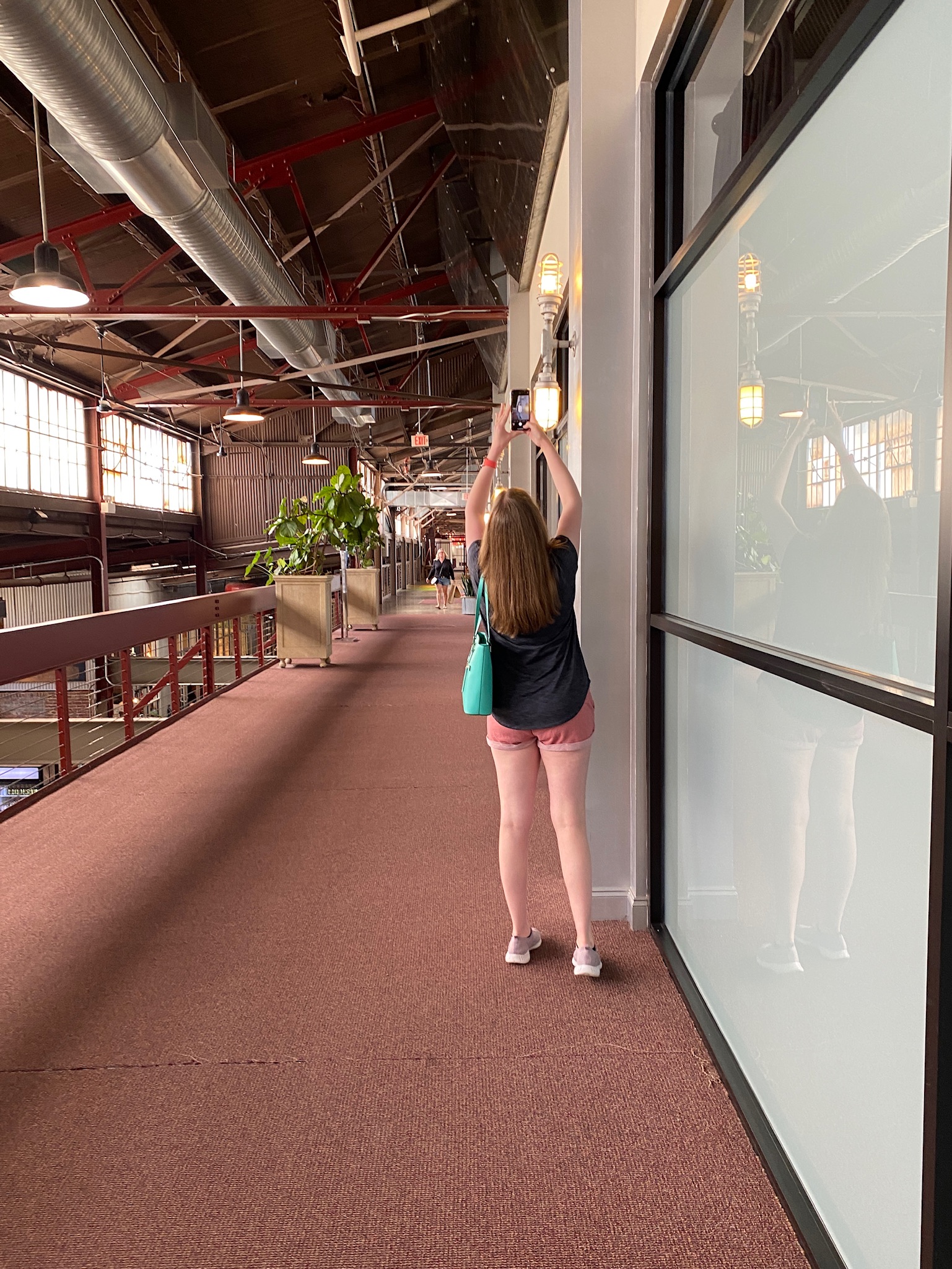 A woman with long hair, wearing a black shirt and pink shorts, stands on a carpeted indoor walkway taking a photo or video with her phone.
