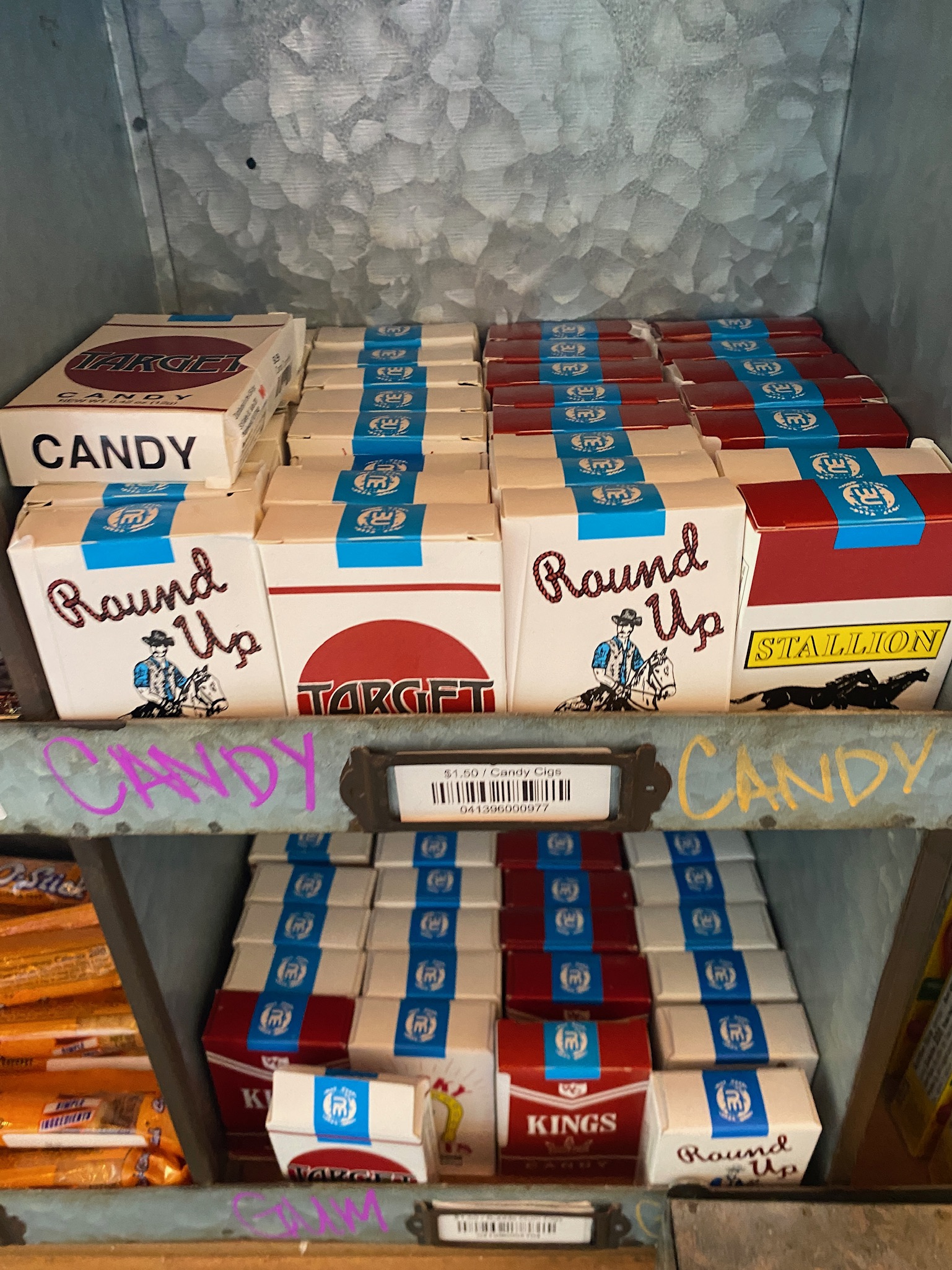 Boxes of candy designed to look like cigarette packs are stacked on shelves labeled “CANDY,” with various brands and colorful packaging visible.