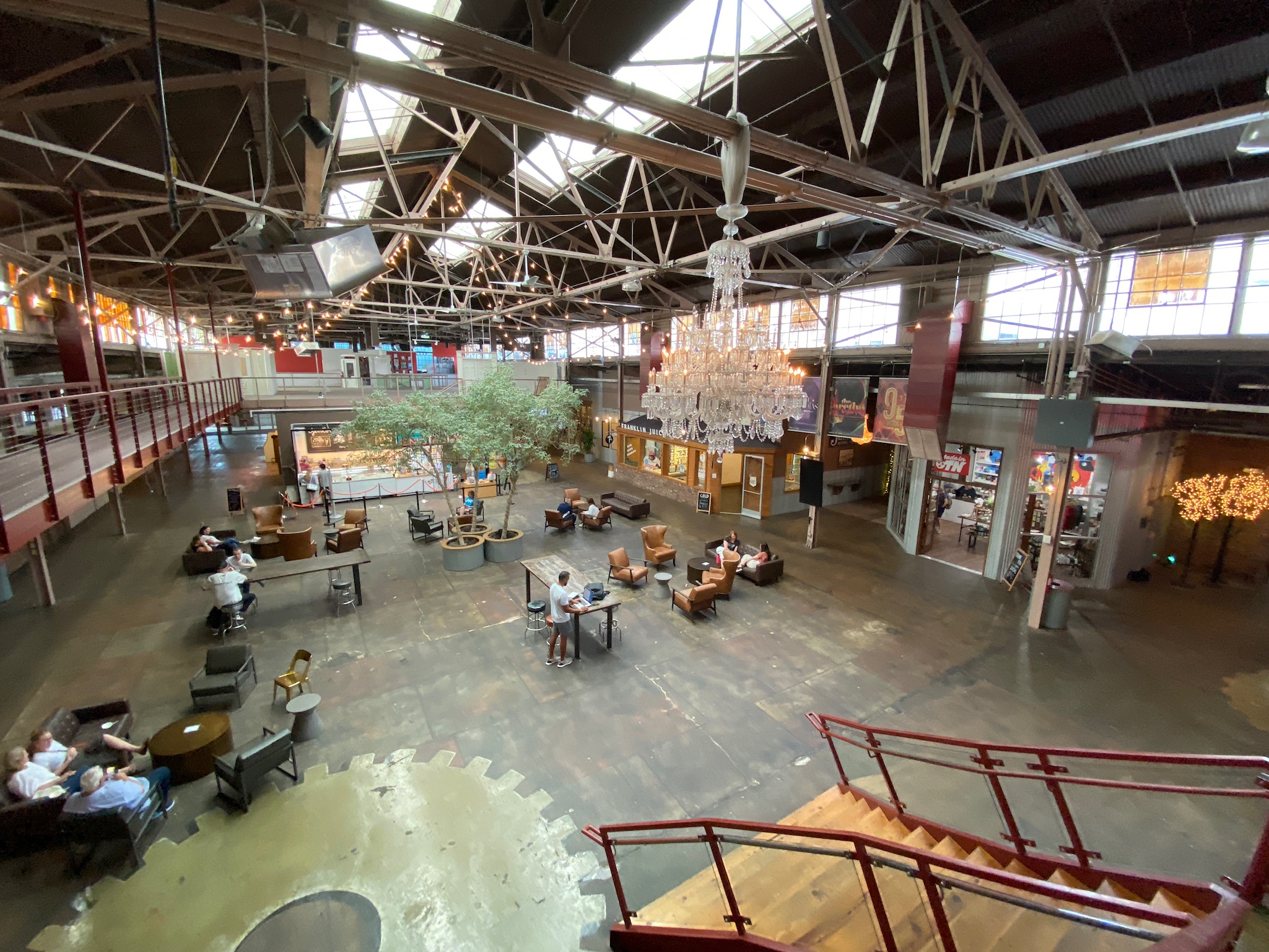 Spacious industrial-style interior with high ceilings, scattered seating, a chandelier, trees, and people sitting at tables; businesses line the perimeter.