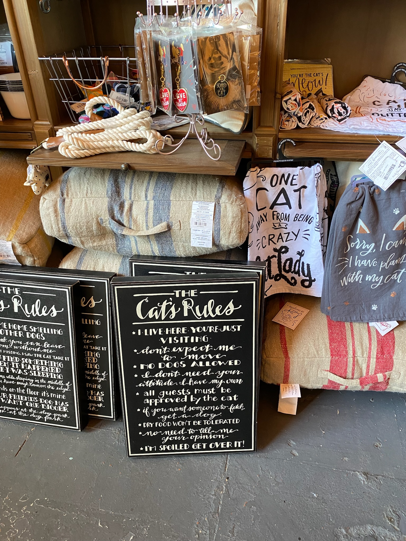 A display of cat-themed home decor items and gifts, including signs, towels, and mugs, with various humorous phrases about cats.