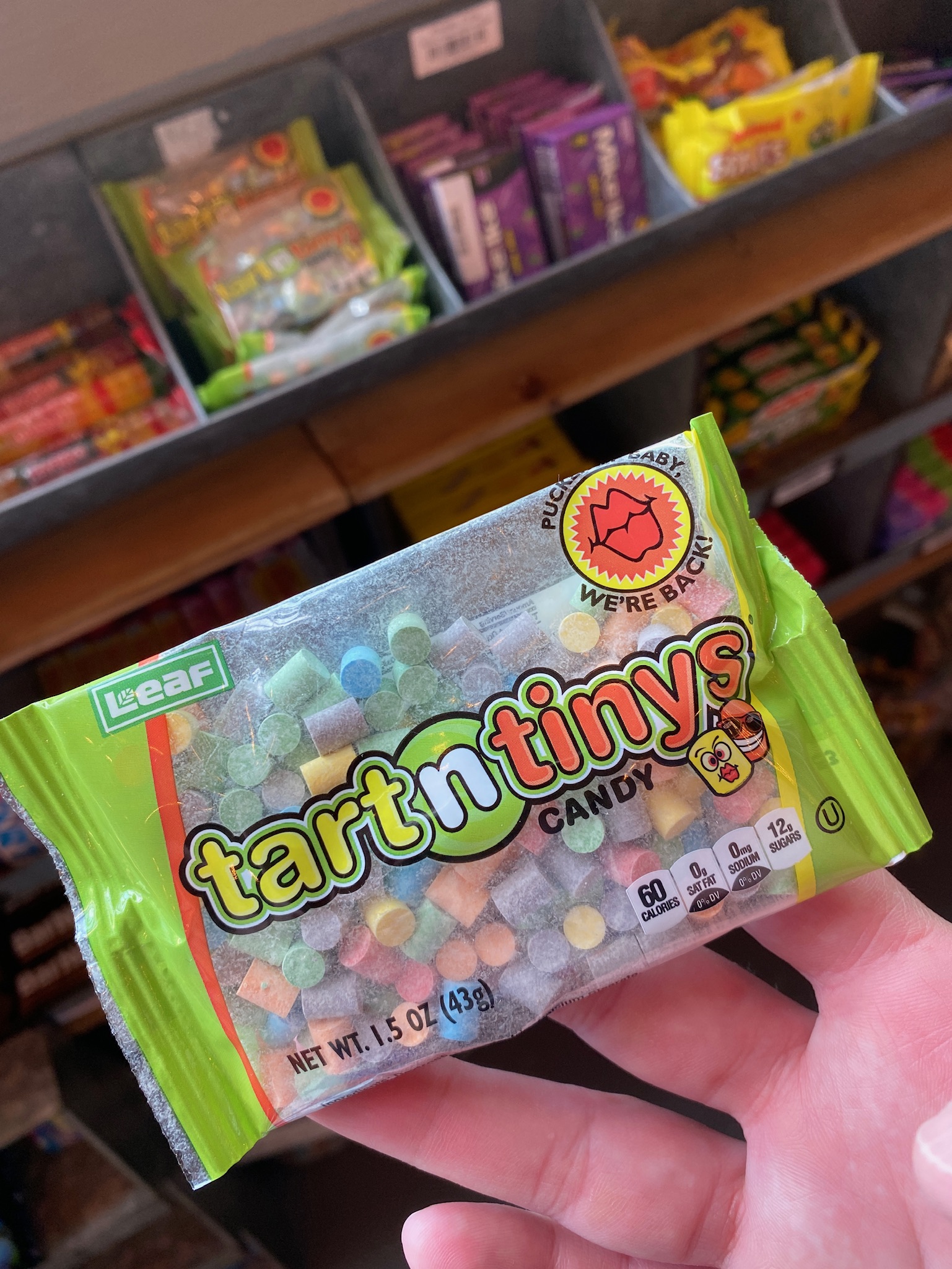 A hand holds a package of tart n tinys candy in front of a shelf stocked with various other candy items.