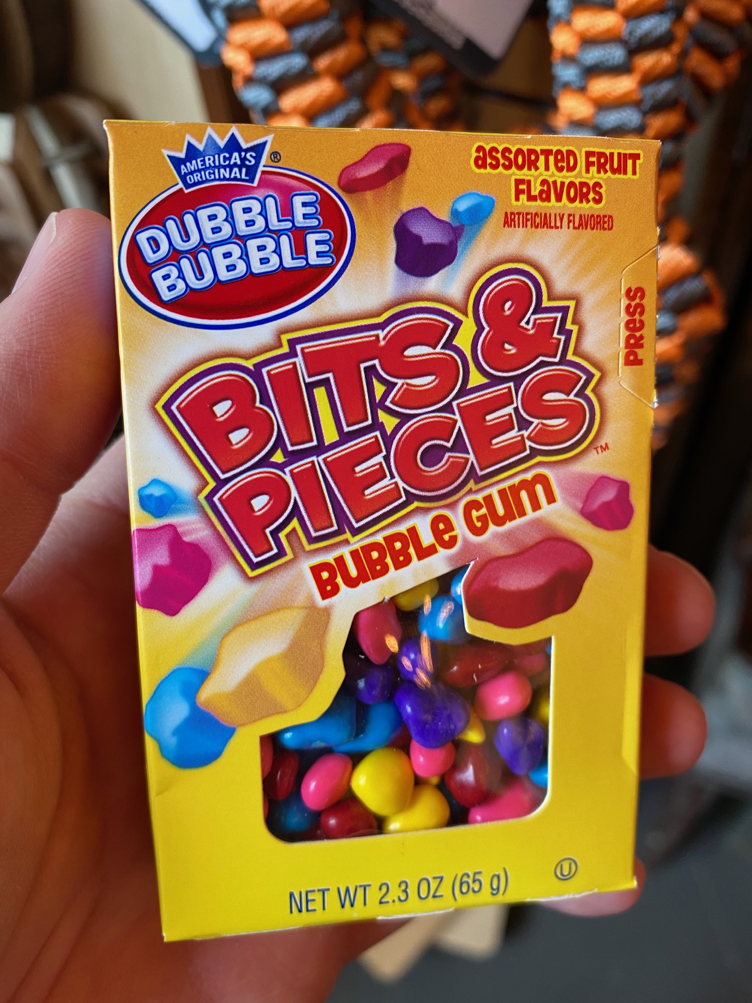 A hand holds a box of Dubble Bubble Bits & Pieces Bubble Gum, assorted fruit flavors, with colorful candy pieces visible through a window in the packaging.